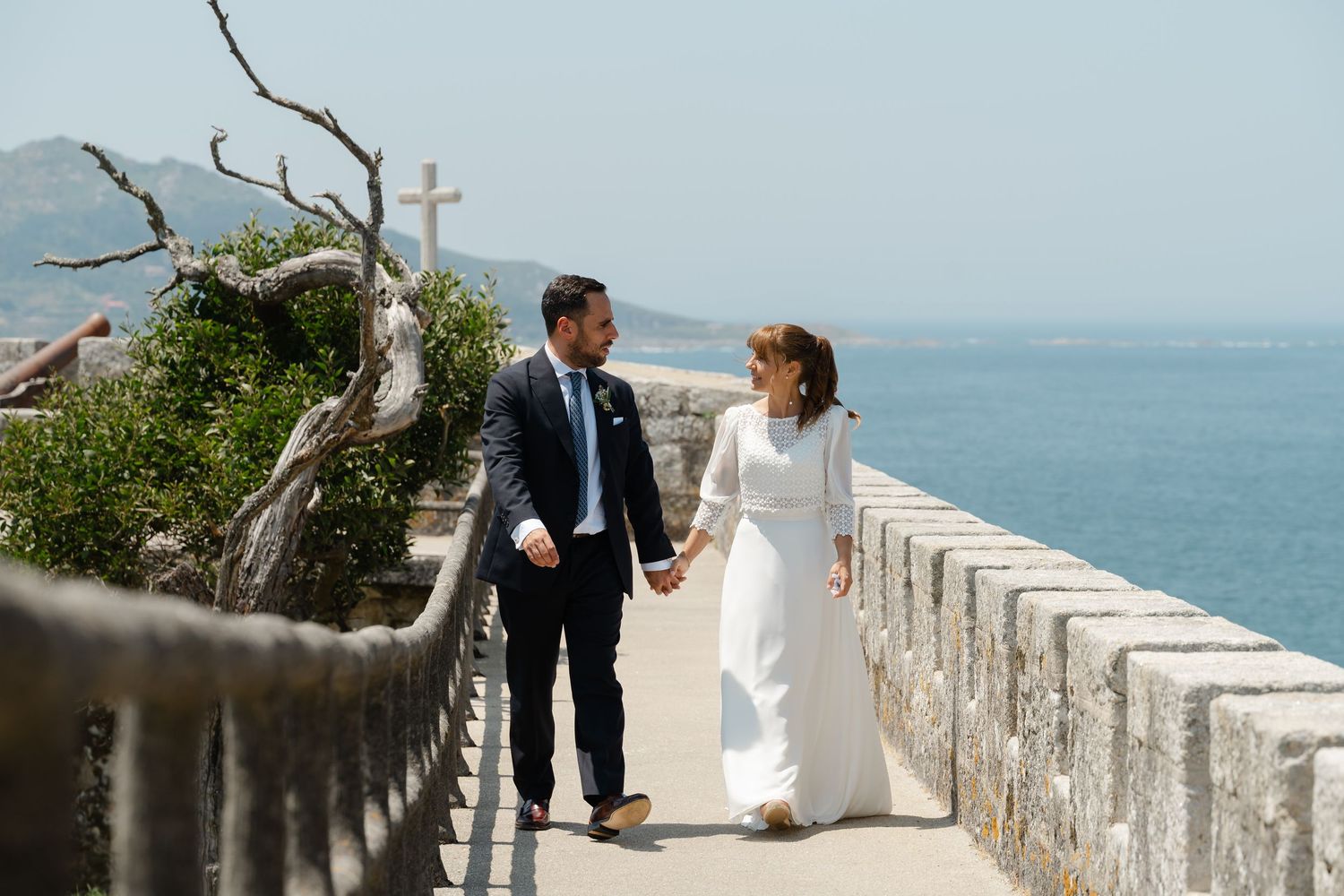 A couple in wedding attire walks along a scenic stone wall overlooking the ocean on a sunny day.