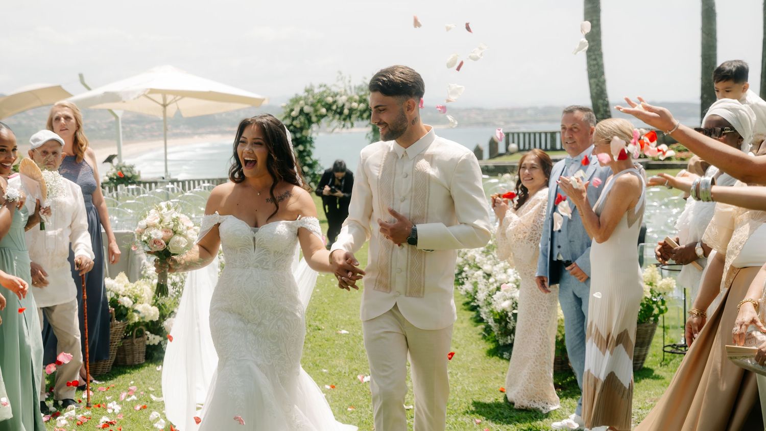 A wedding party celebrates outdoors with guests in white and pastel attire during a sunny daytime reception.