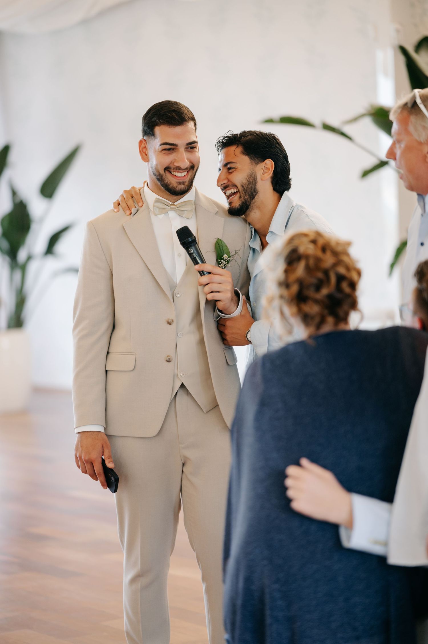 Wedding guests share joyful moments and laughter during a bright indoor celebration with tropical plants.