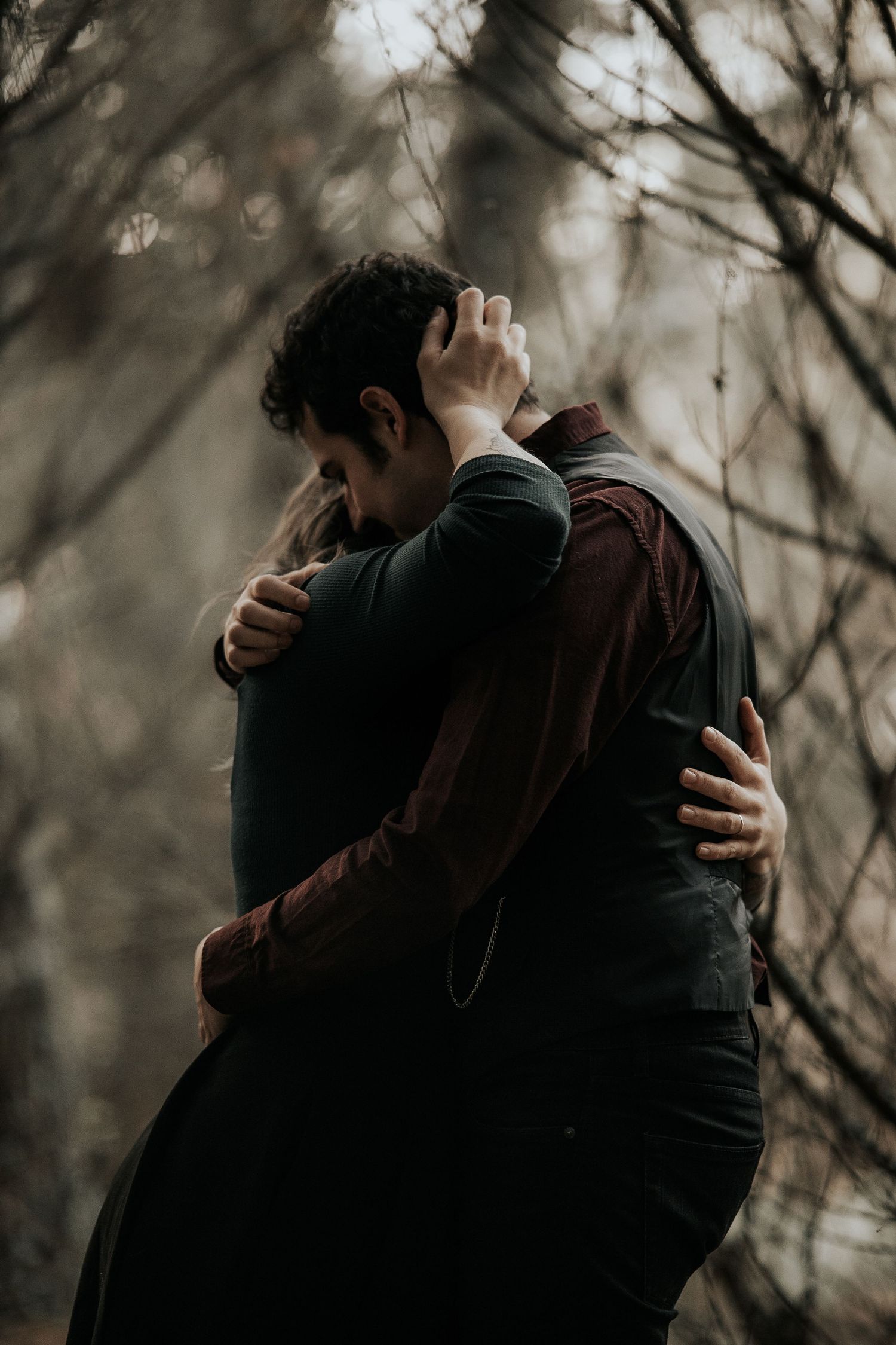 A romantic couple embraces intimately in a moody forest setting with bare winter trees in the background.