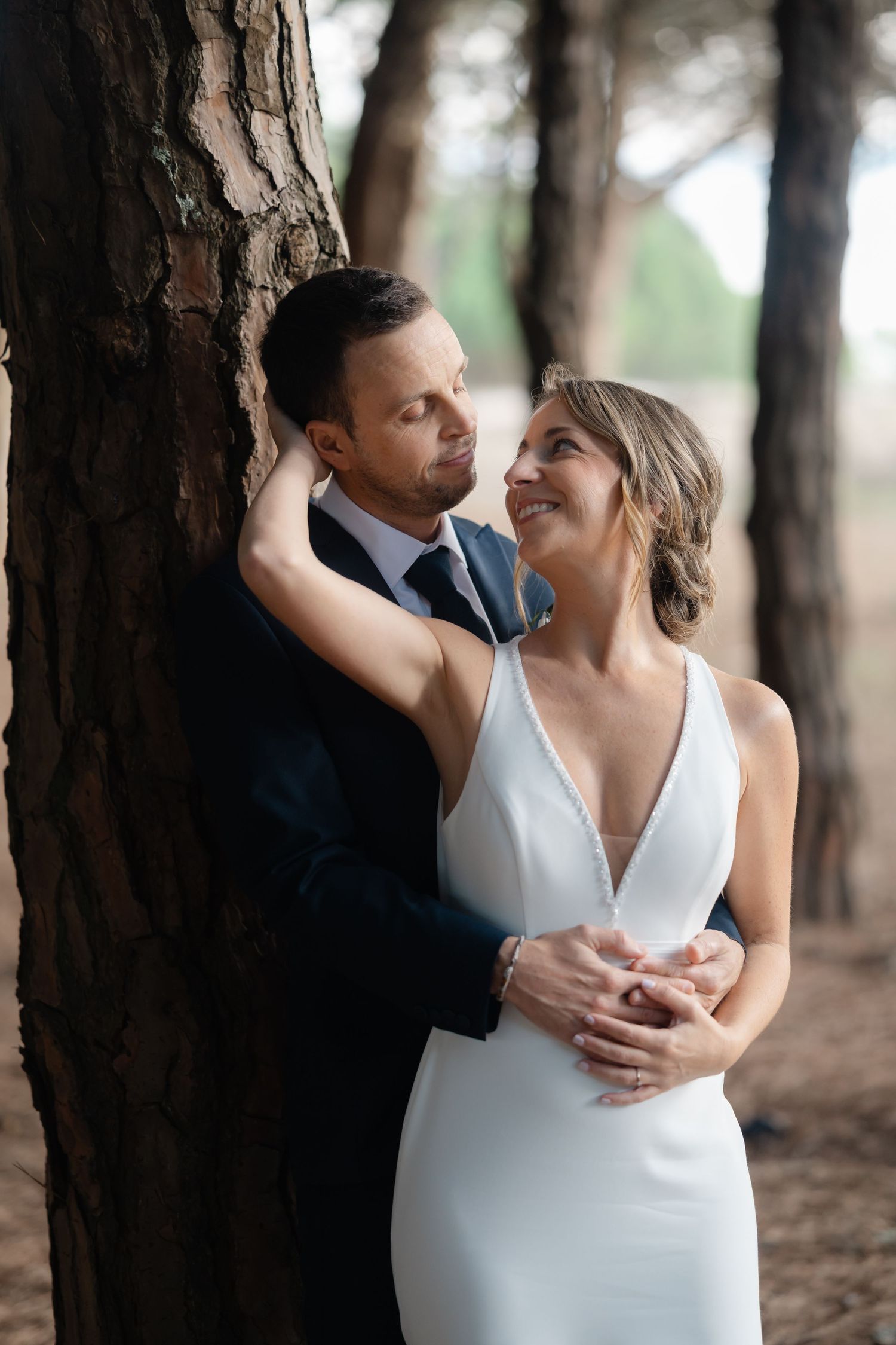 A romantic wedding couple embraces against a tree trunk in a serene forest setting.