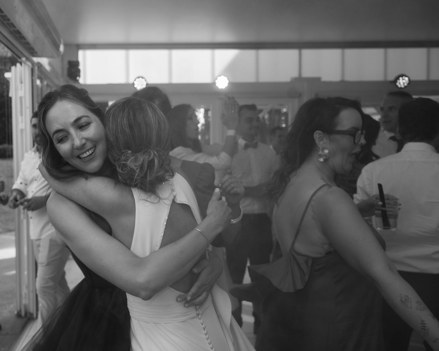 Two people embrace in a warm hug while dancing at a social gathering, with other guests visible in the background.
