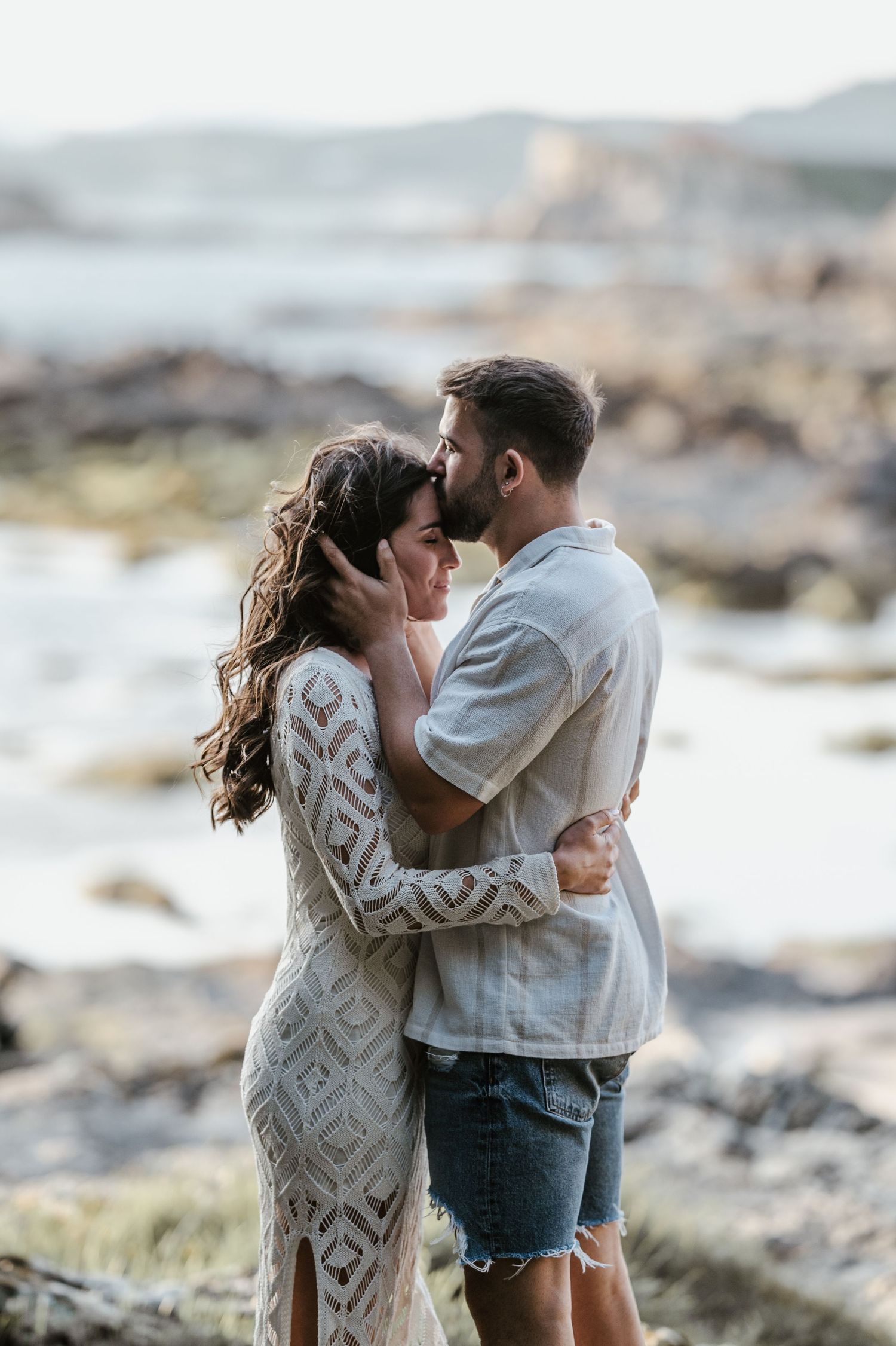 Couple embracing on a rocky beach shoreline at sunset wearing white and denim casual outfits.