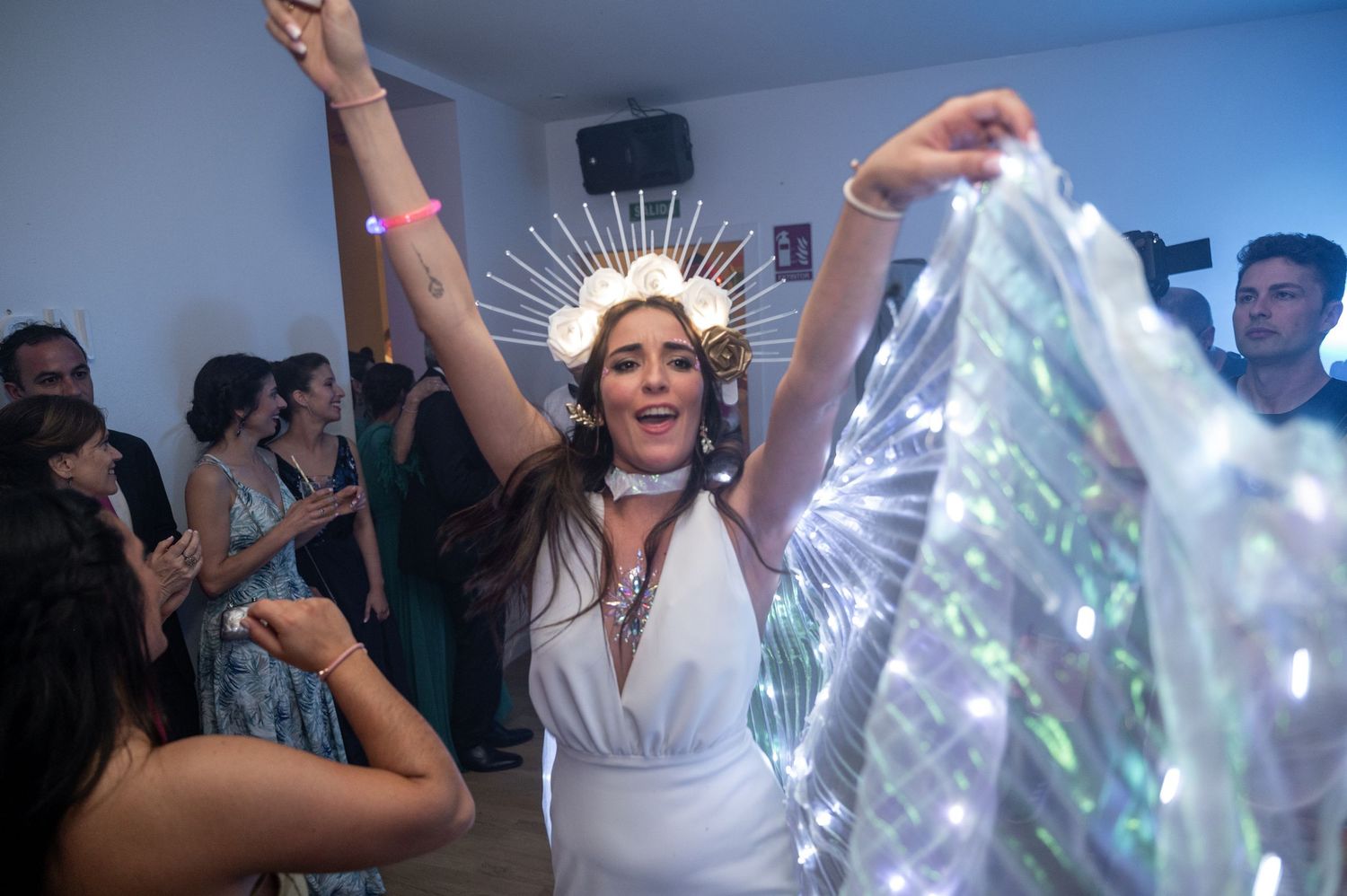 A woman in white dress and crown dancing joyfully at a party with raised arms and shimmering decorations.