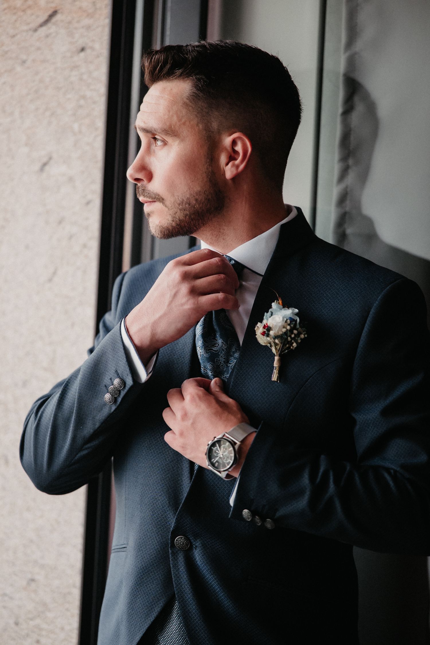 Groom in navy suit adjusts his tie while looking out window in contemplative wedding day portrait series.