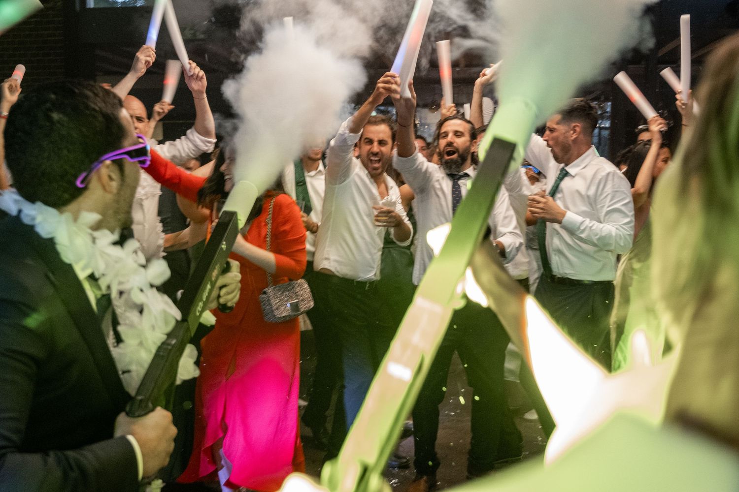 Wedding celebration with guests raising glasses through atmospheric smoke and colored lighting effects.