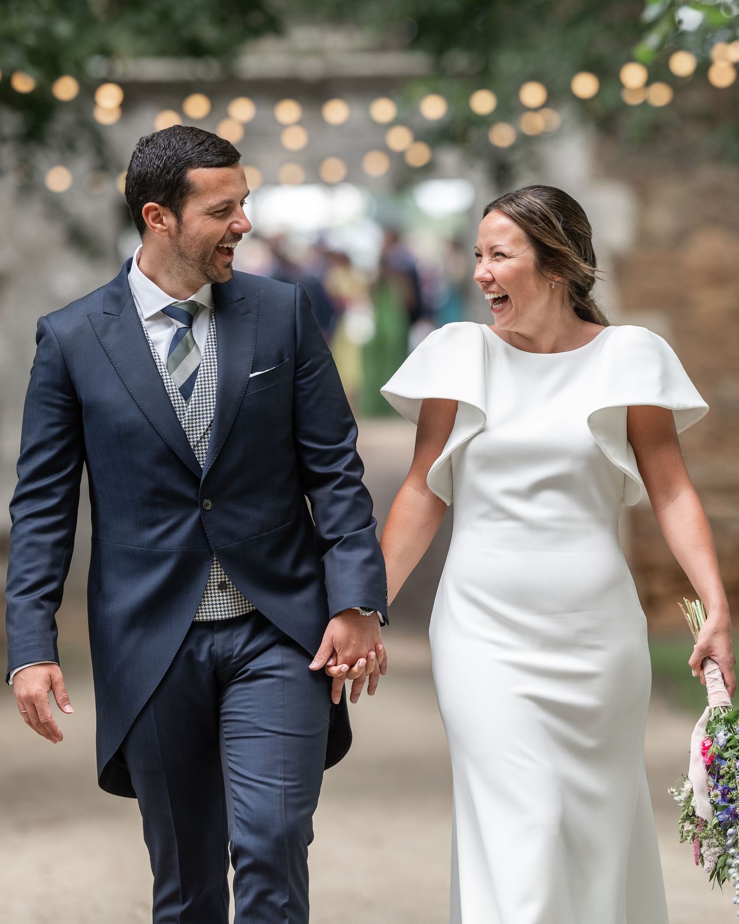 A happy couple in wedding attire walking and laughing together under string lights in an outdoor setting.