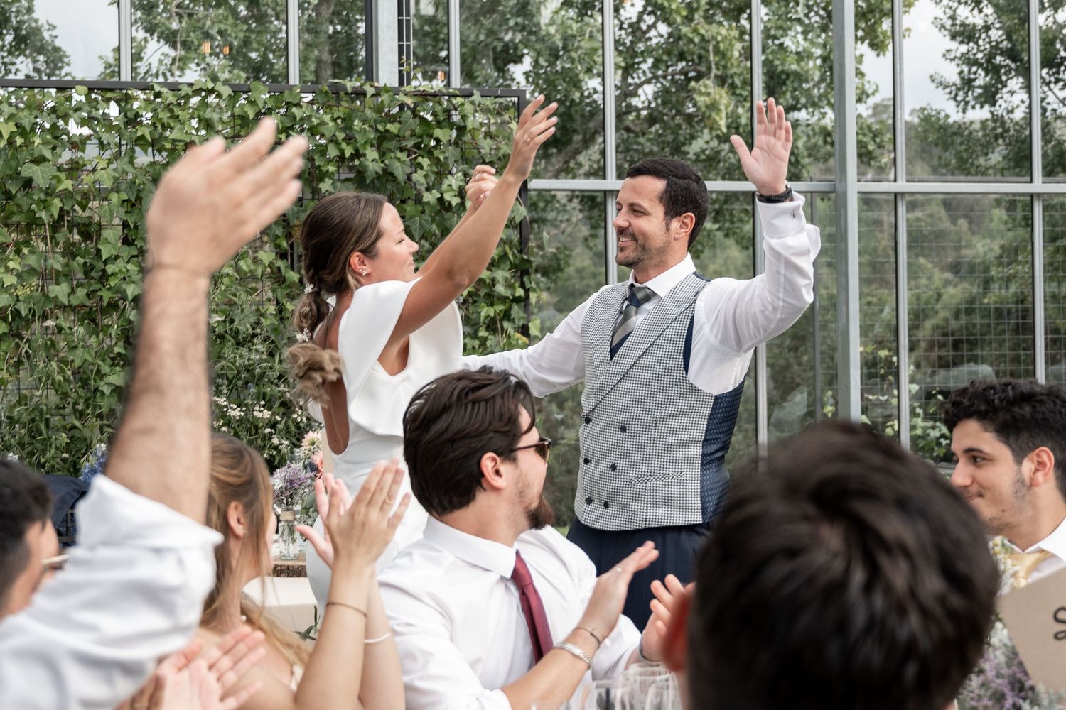 Wedding guests raise their arms in celebration during an outdoor reception with trees in the background.
