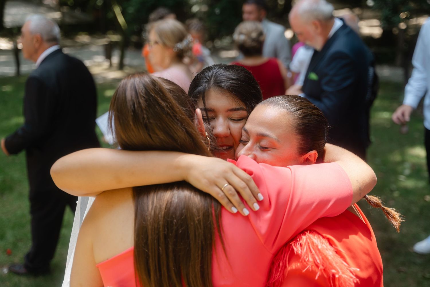 Wedding guests embrace in warm hugs while wearing bright coral colored attire at an outdoor celebration.