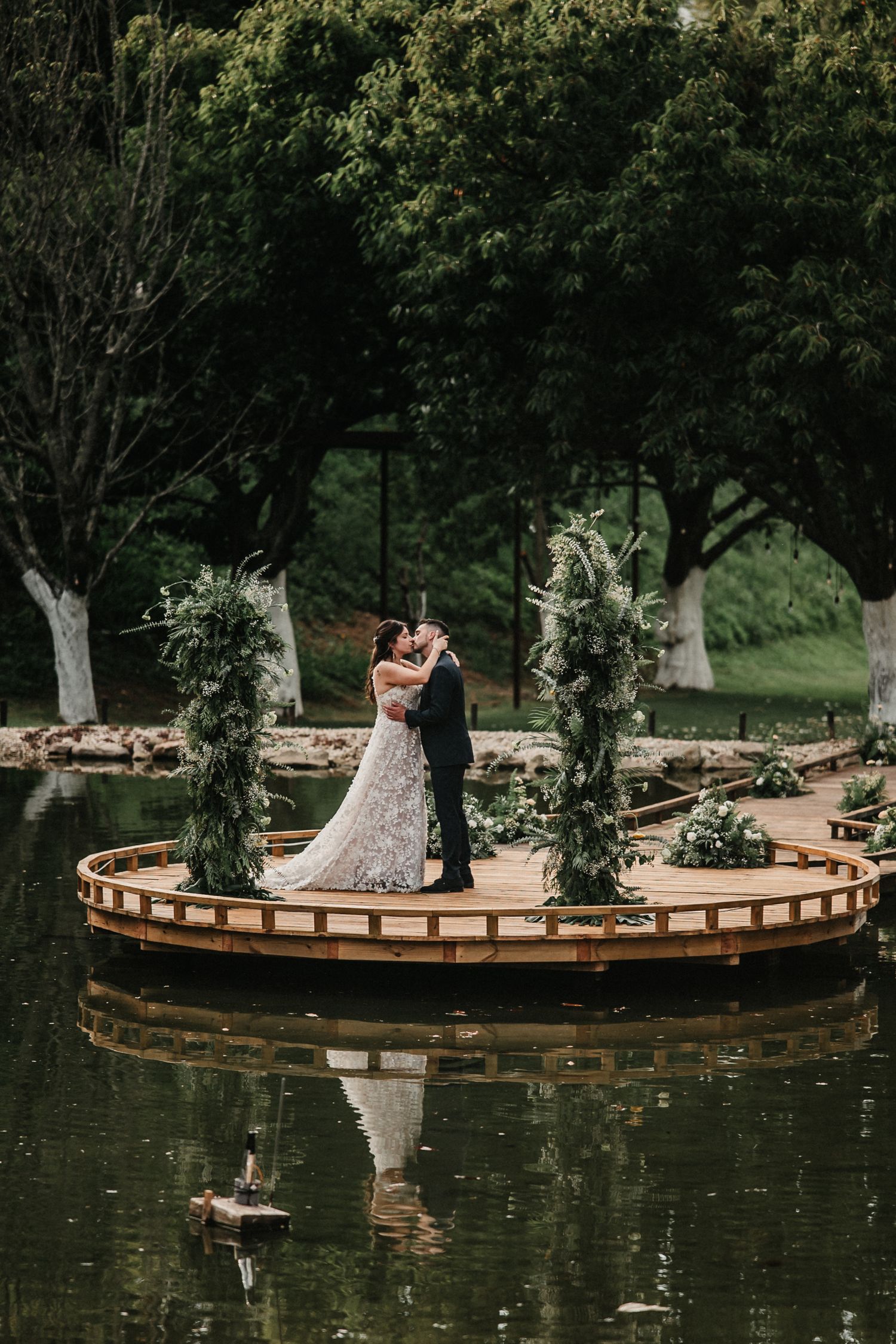 Circular wooden deck extends over a serene pond surrounded by lush trees and greenery in a tranquil garden setting.