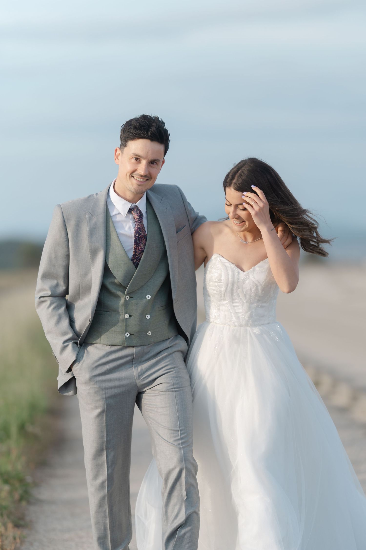 A smiling bride and groom walk together on a beach path, she in a white gown, he in a gray suit.