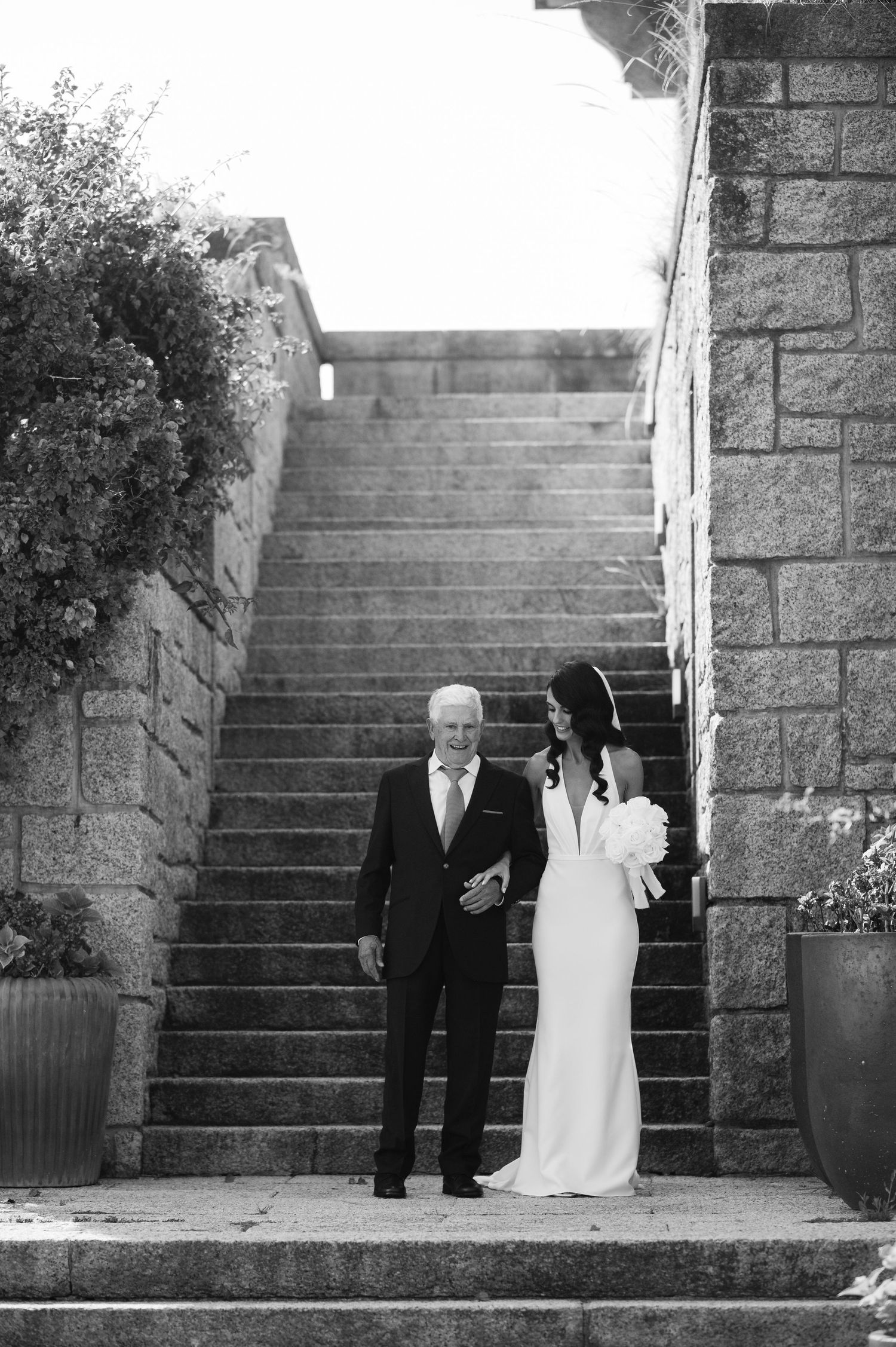 Black and white photo of a bride in a fitted white dress and companion walking down stone steps at an outdoor venue.