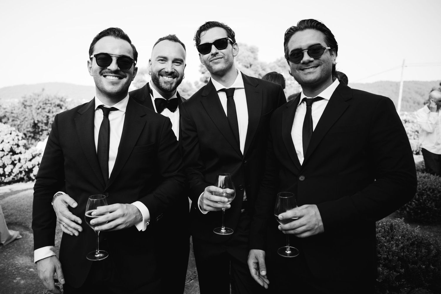 Groomsmen in black suits and sunglasses pose with drinks outdoors at a wedding celebration.