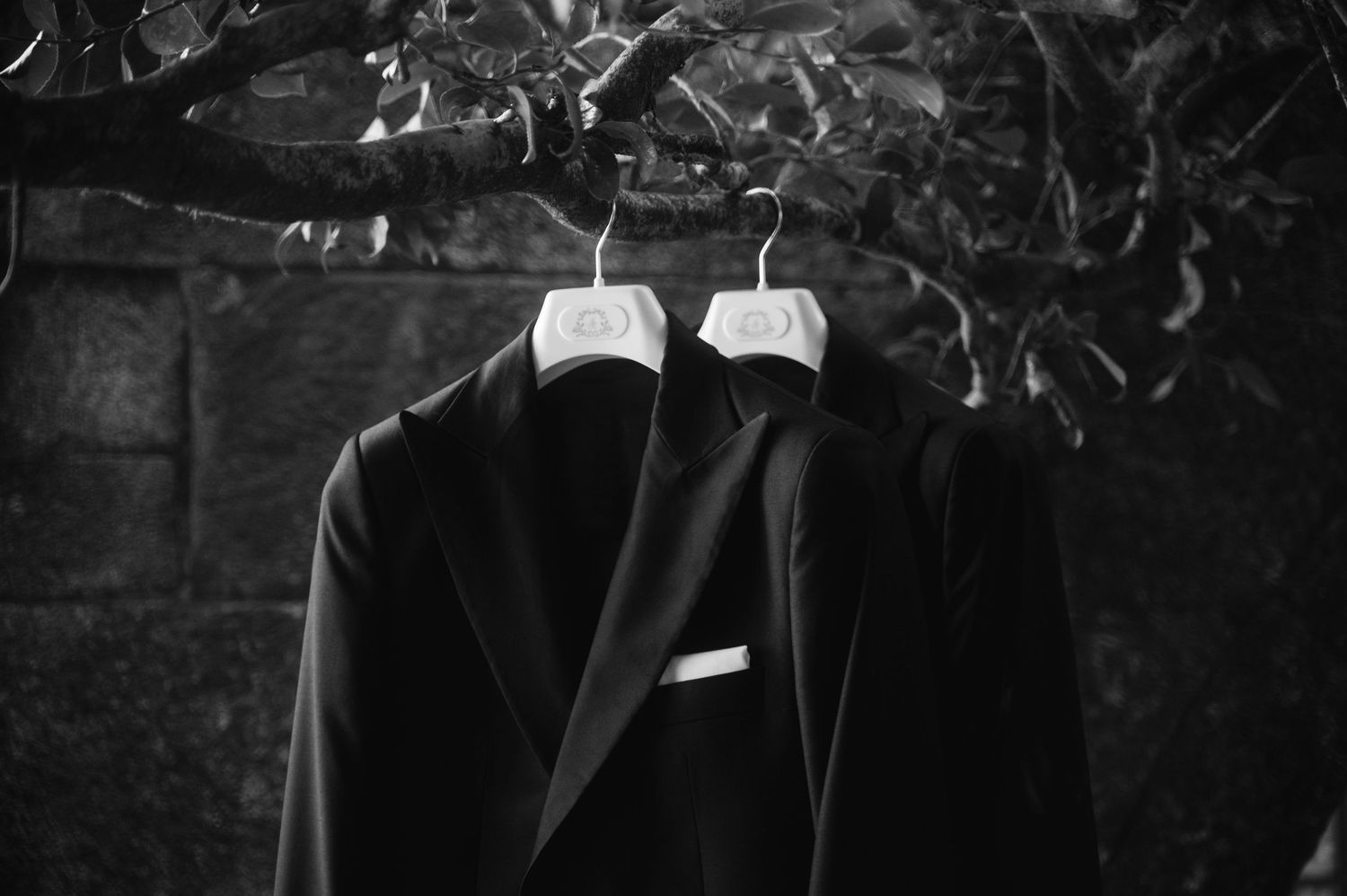 Two black formal tuxedo jackets hanging on white hangers against a dark textured background.