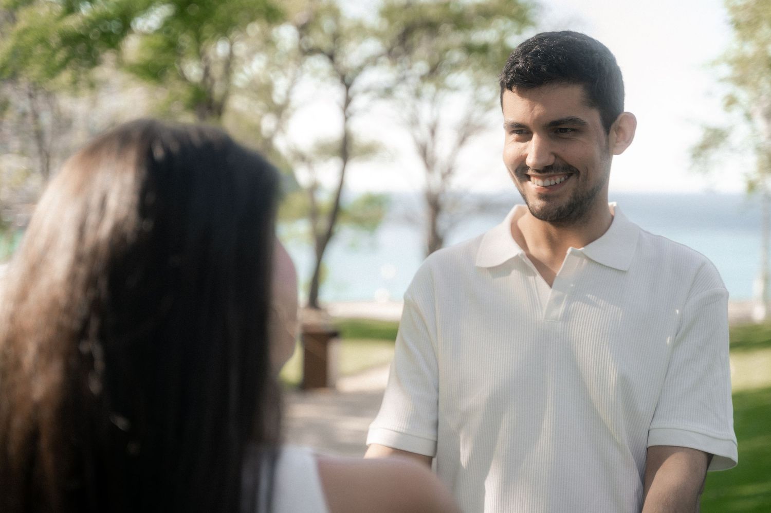 A couple in white outfits walks along a scenic lakeside path during a romantic outdoor photoshoot.