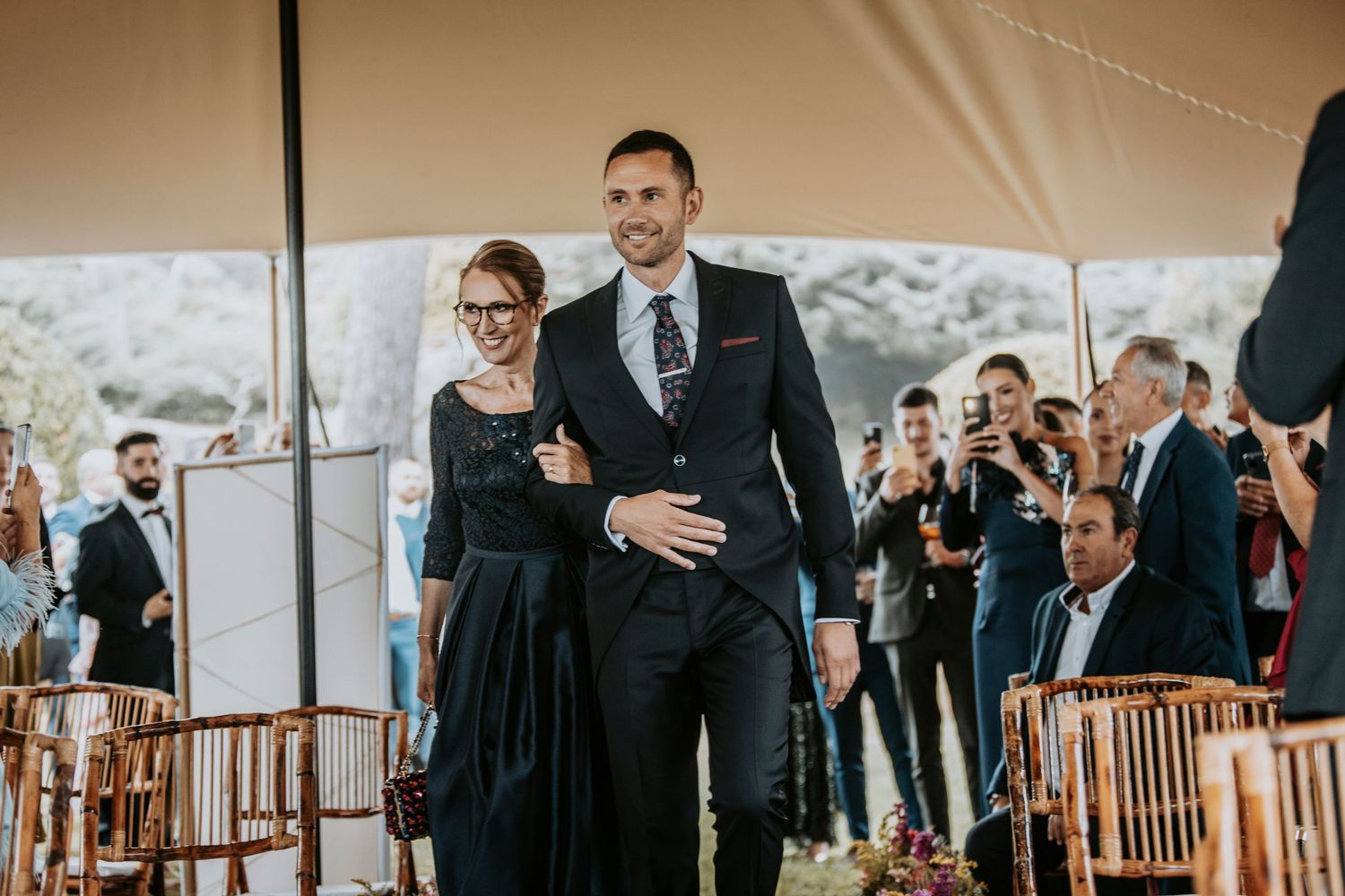 A couple stands together during a wedding ceremony while guests look on under a white tent.