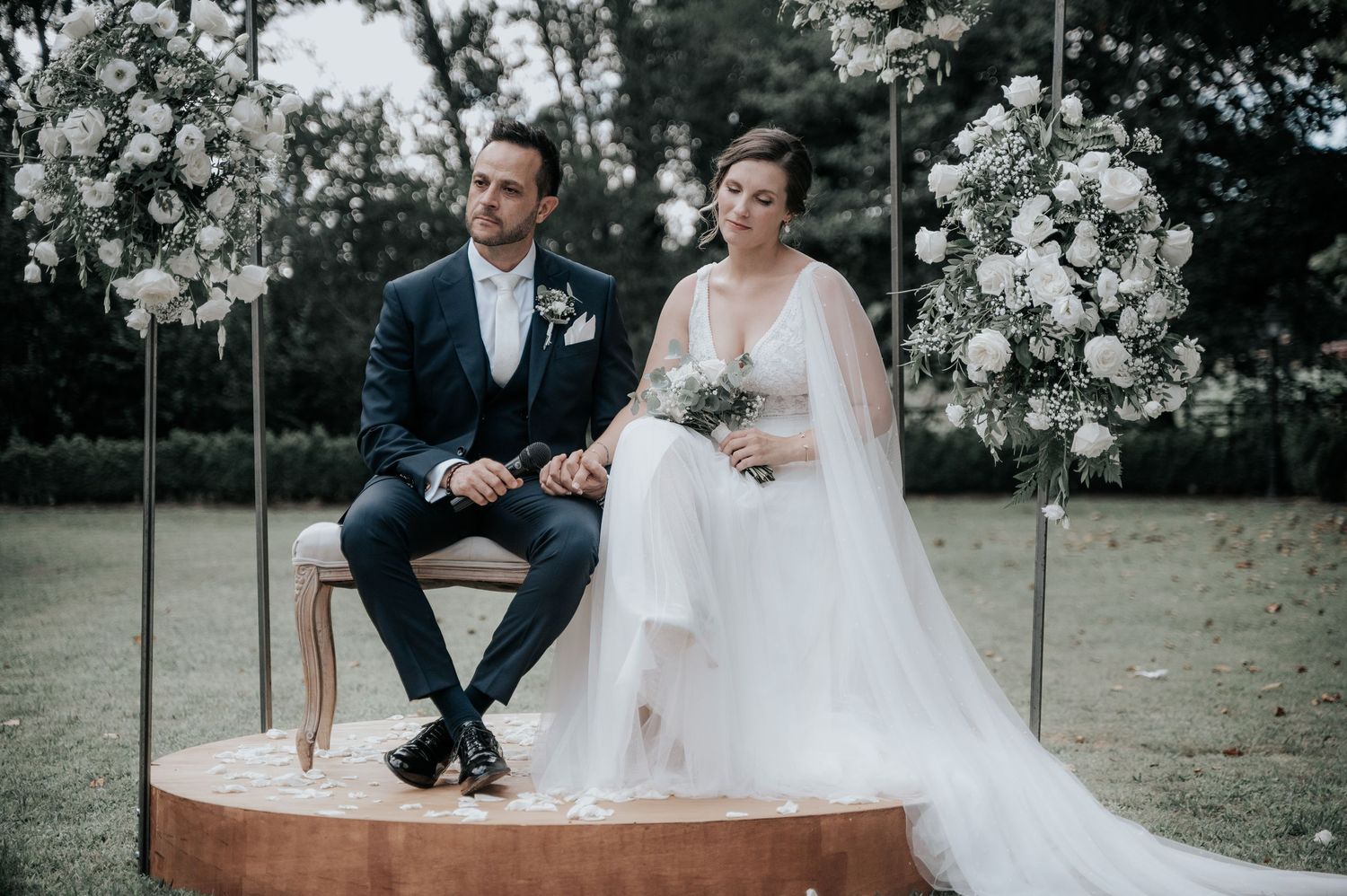 A couple sits together on a rustic white fountain in an outdoor garden setting during a wedding celebration.
