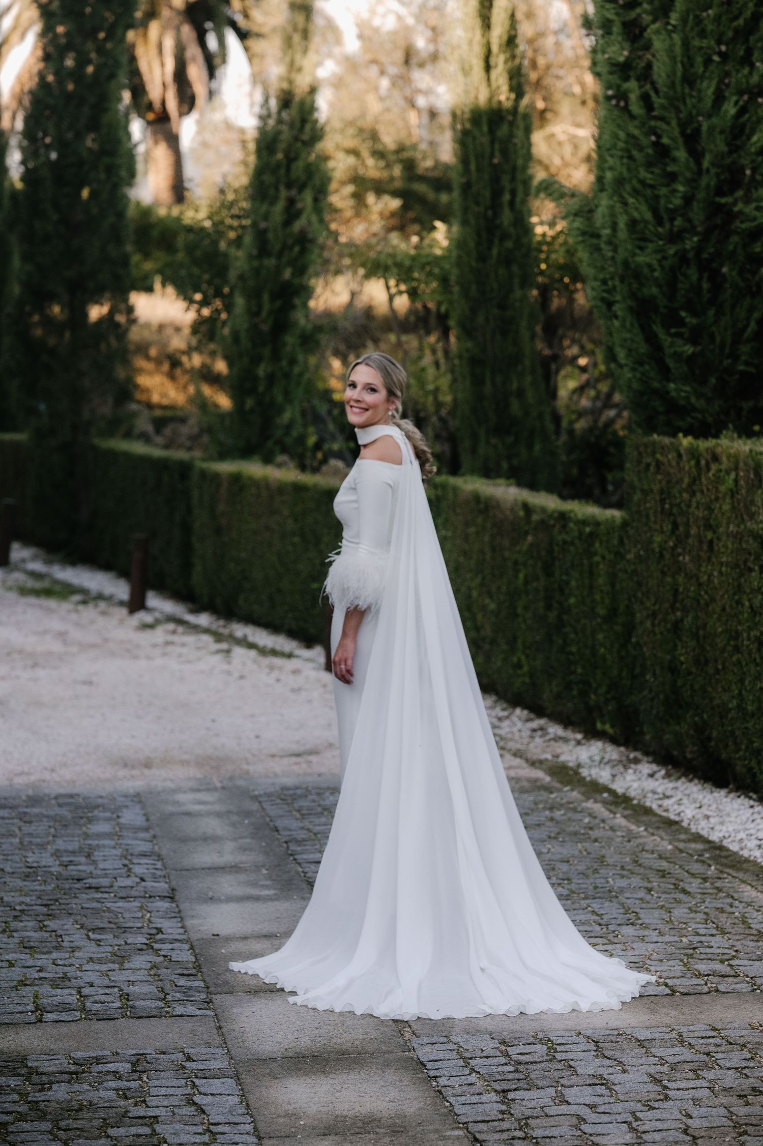 Elegant bride in white gown with flowing cape walking along cobblestone path surrounded by manicured hedges.
