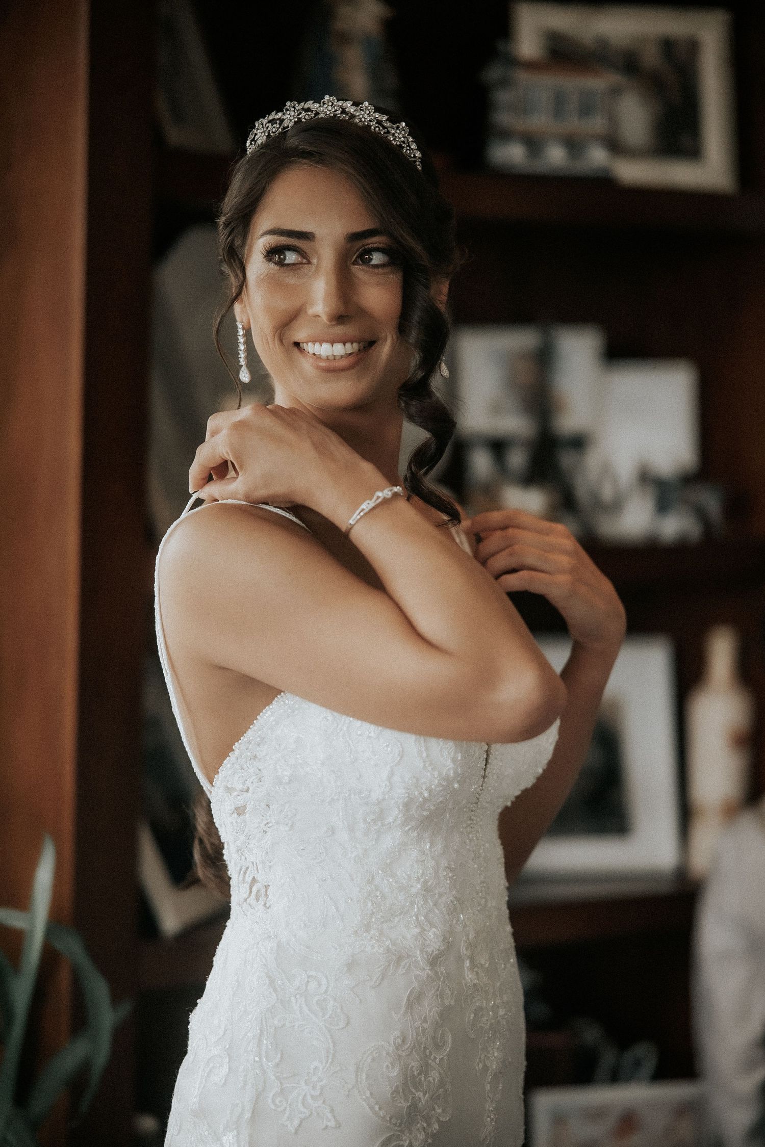 A bride in a stunning white lace gown and delicate tiara poses elegantly against a dark background.