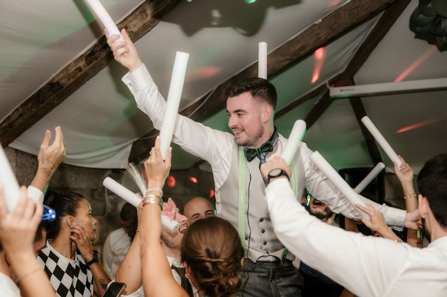 People dancing and celebrating with LED foam sticks at a lively party under a white tent.
