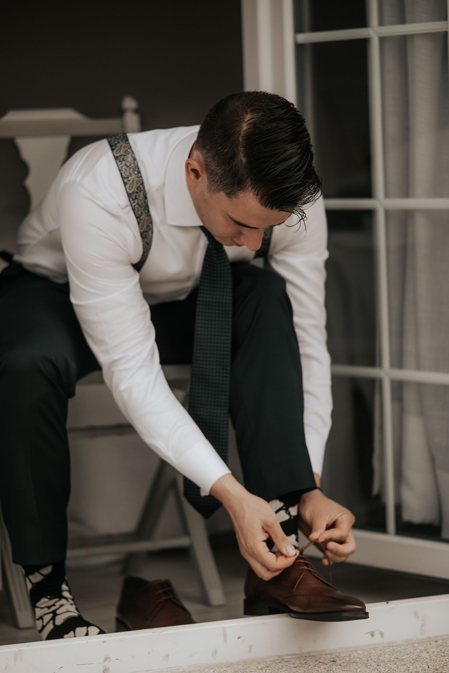A person in formal attire ties brown leather dress shoes while getting ready for a special occasion.