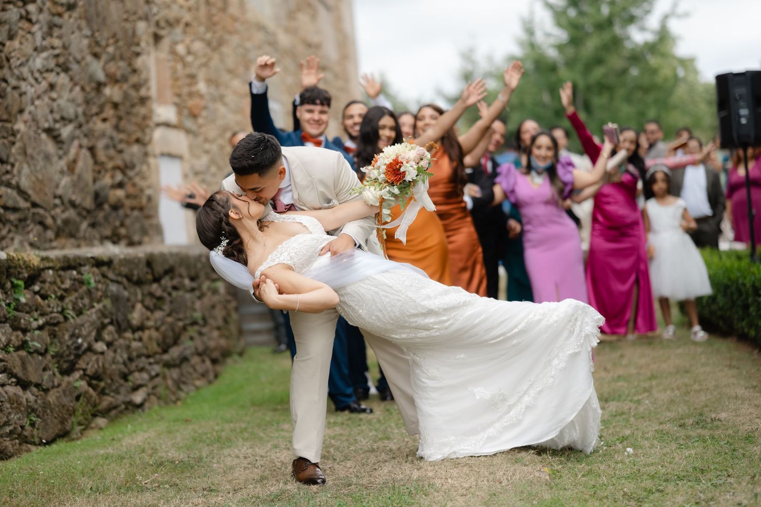 Wedding party celebrates with dramatic dip poses on grassy lawn near stone wall.