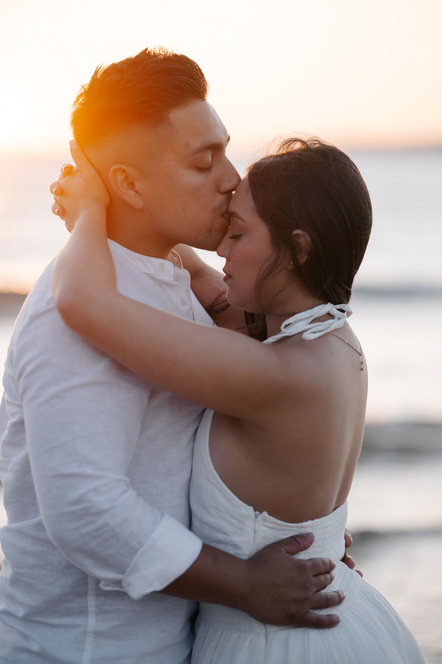 Romantic sunset beach photoshoot with couple embracing in white outfits.