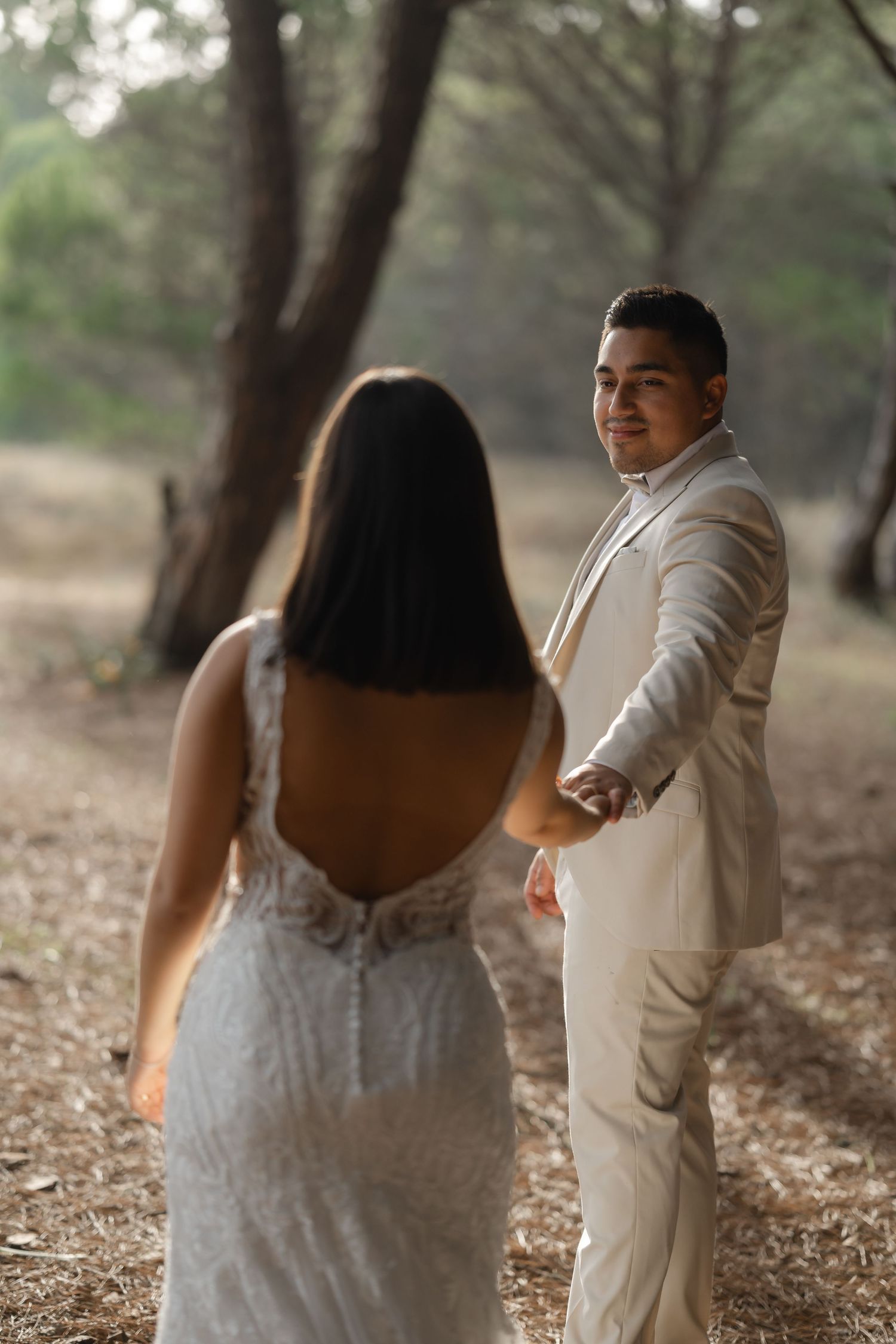 Romantic wedding couple in white dress and beige suit embrace in a wooded outdoor setting.