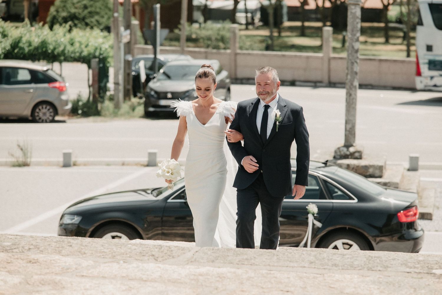 A couple in formal attire walks together outdoors near parked cars on a bright day.