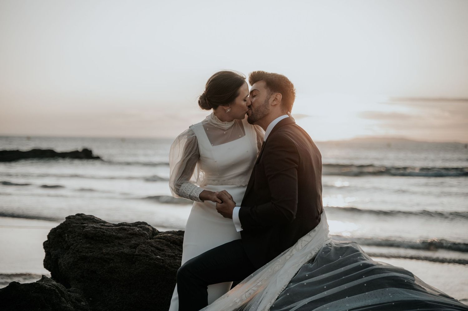 Couple embracing on rocky beach at sunset with ocean waves in background.