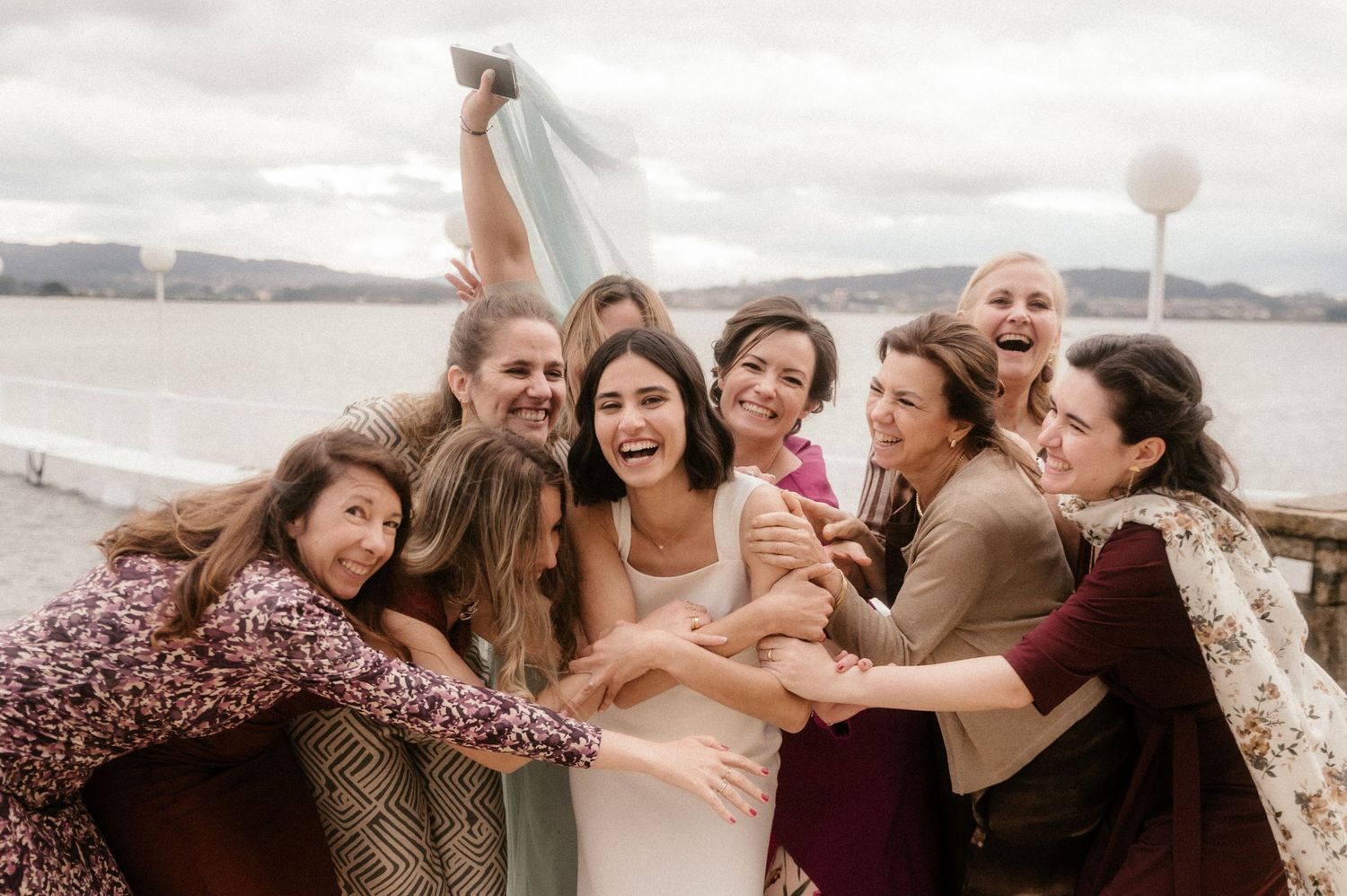 A group of friends huddle together and laugh joyfully on a cloudy day by the water.