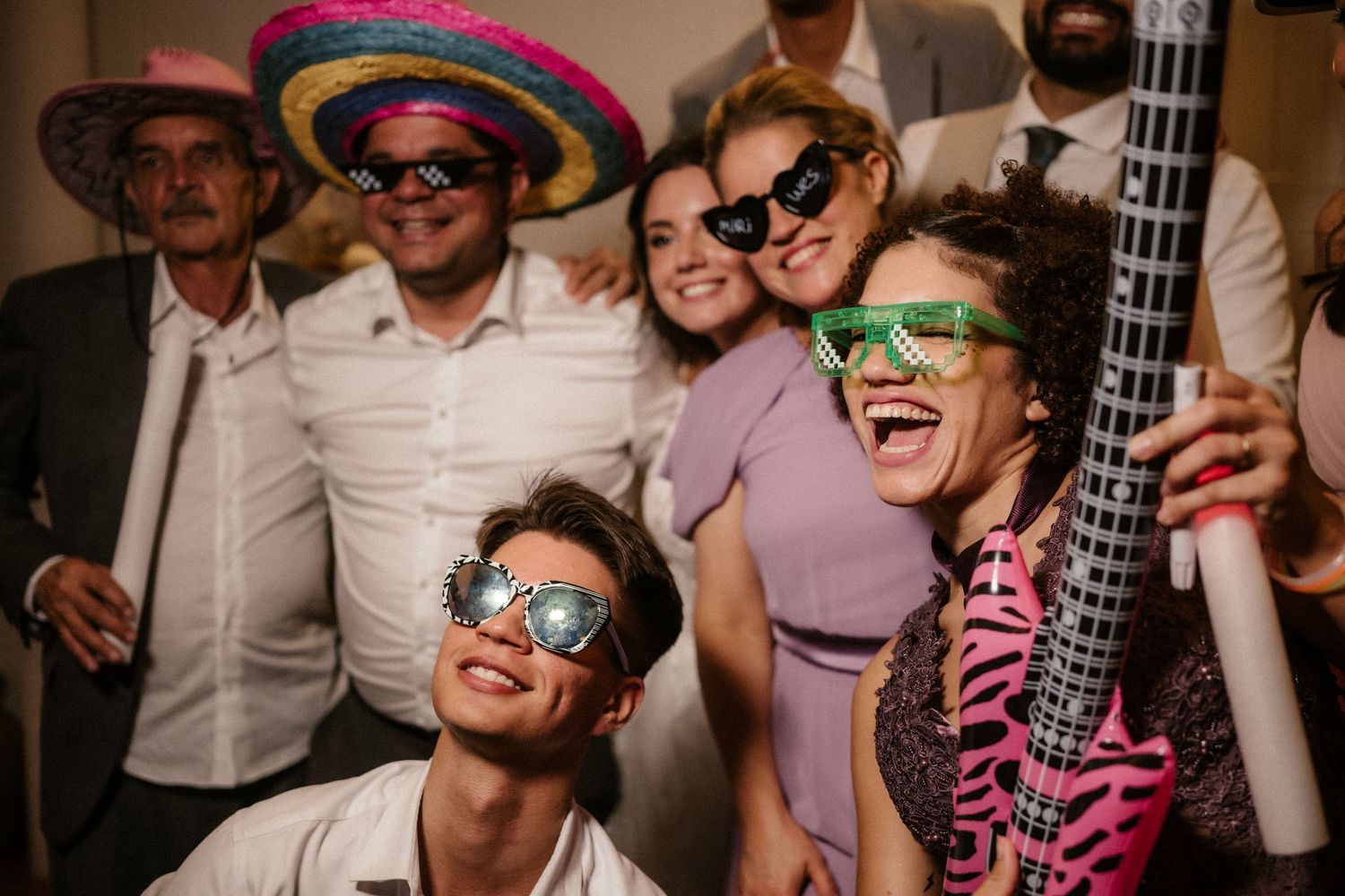 Wedding party guests wearing colorful accessories and sunglasses dance and celebrate on the dance floor.