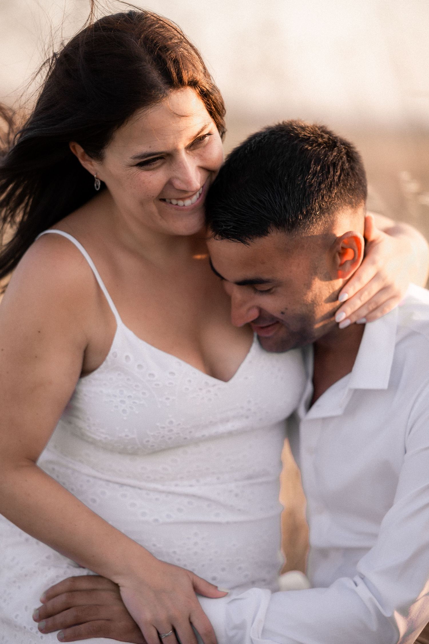 Intimate candid moment of couple laughing and embracing in white outfits during sunset photoshoot.