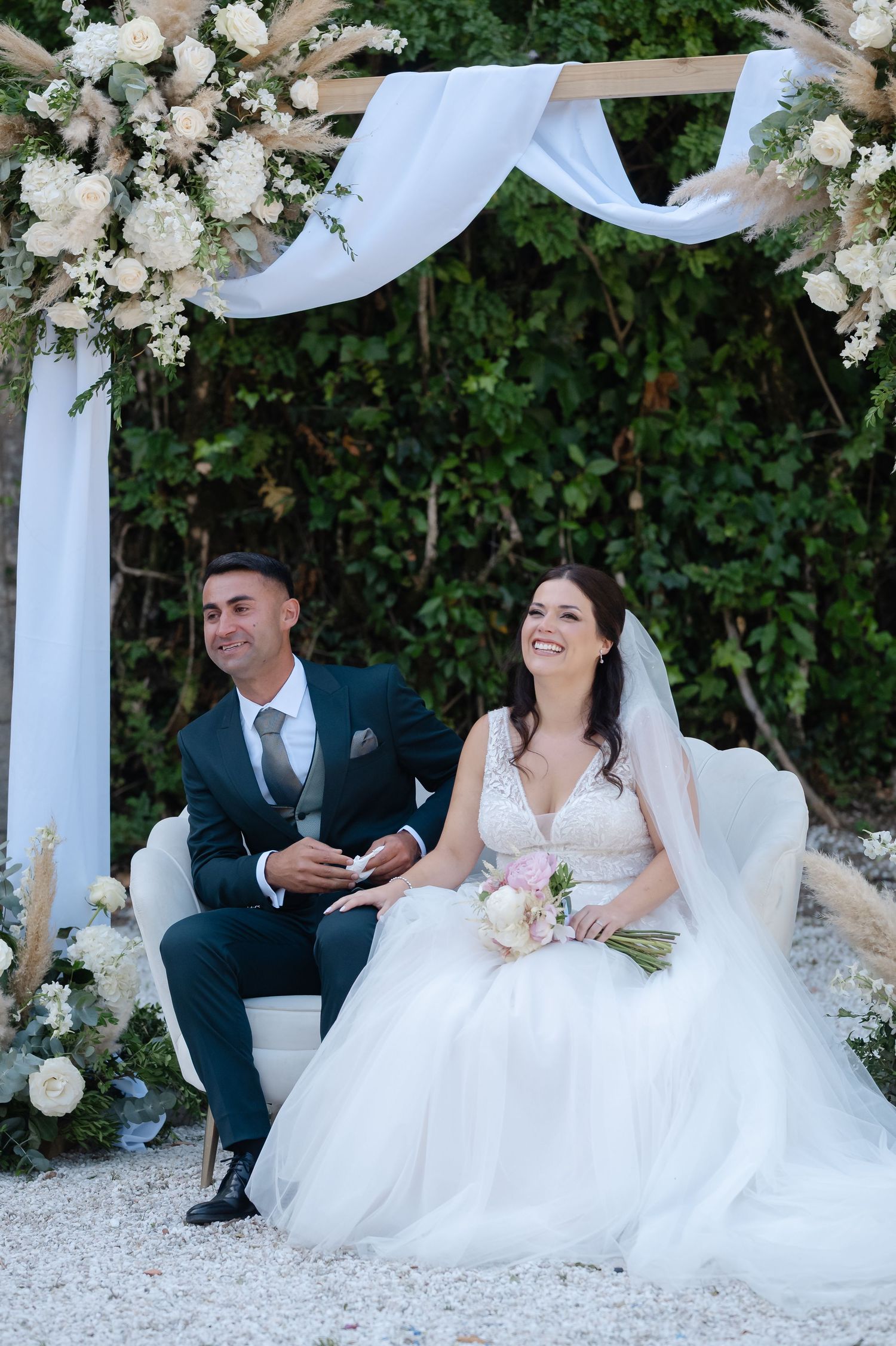 Wedding party sits on white chairs under an elegant floral arch decorated with white flowers and flowing fabric.