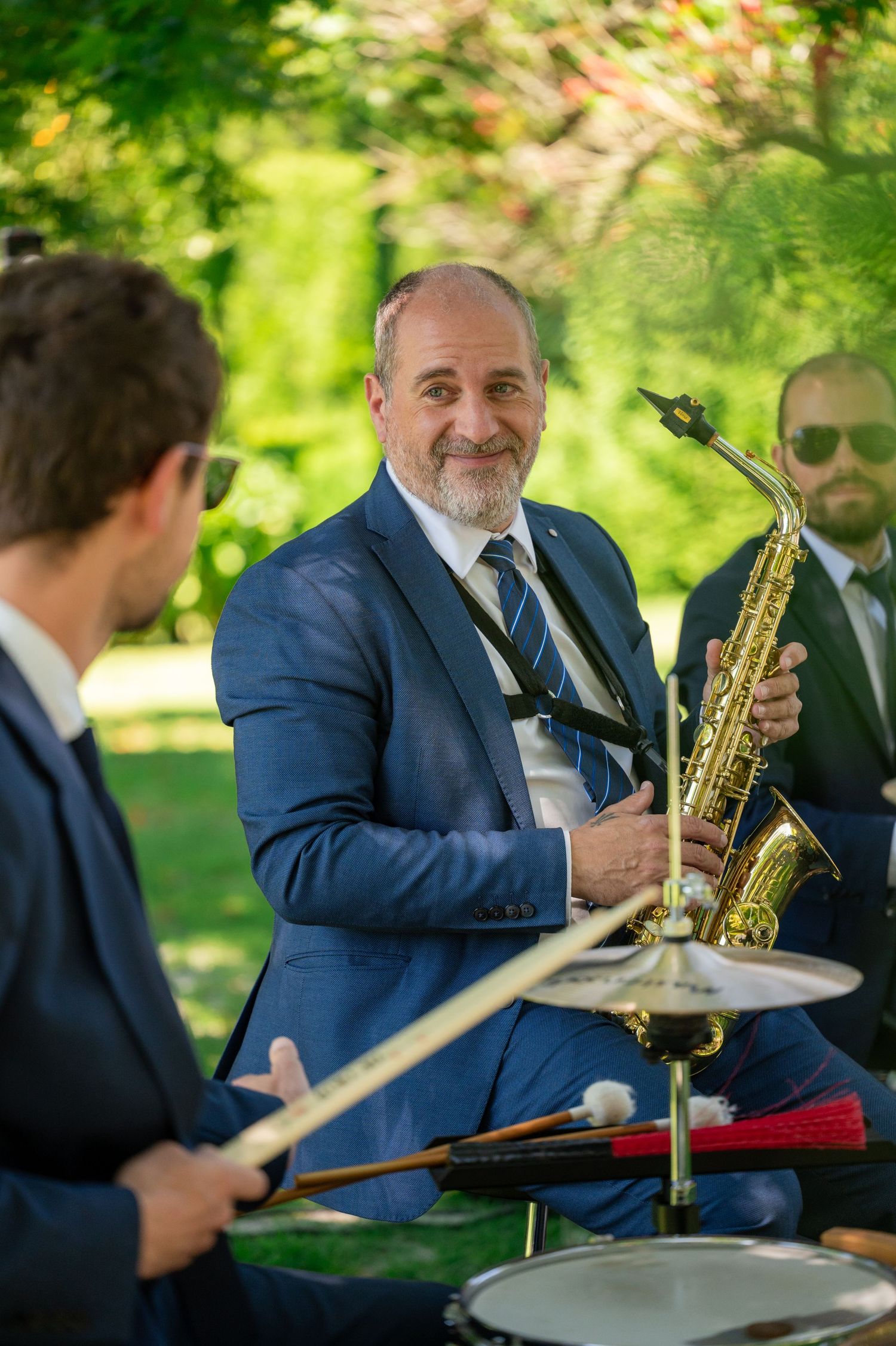 Musicians performing outdoors with instruments including saxophone and drums in a sunny garden setting.