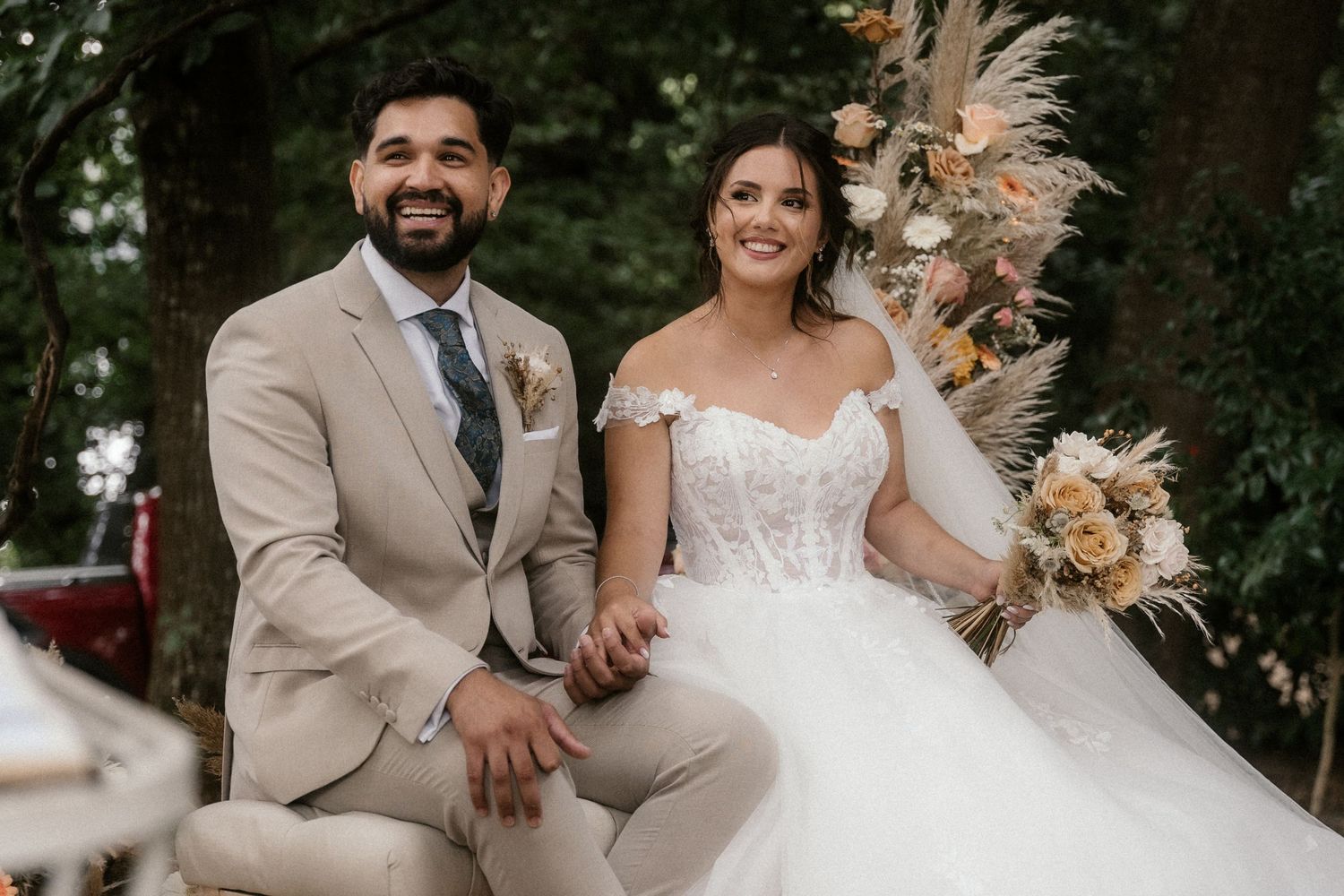 Wedding party poses together outdoors wearing beige suits and white dresses against natural backdrop.