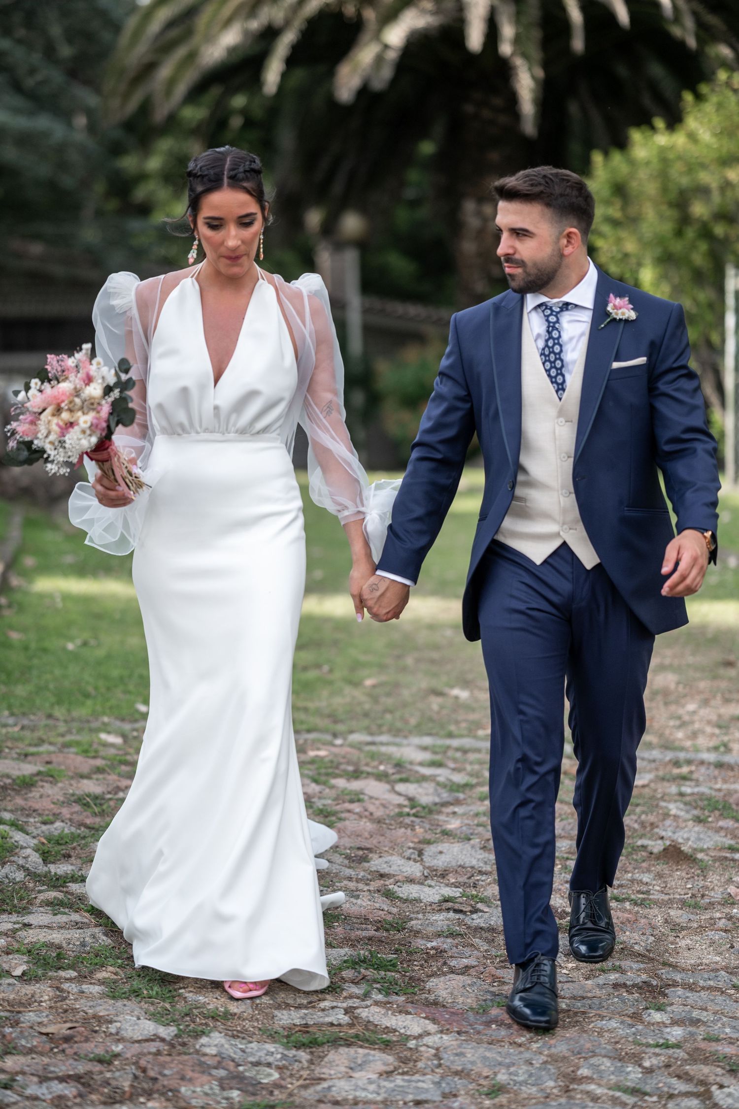 Newlywed couple walks together on a cobblestone path with bride in white dress and groom in navy suit.