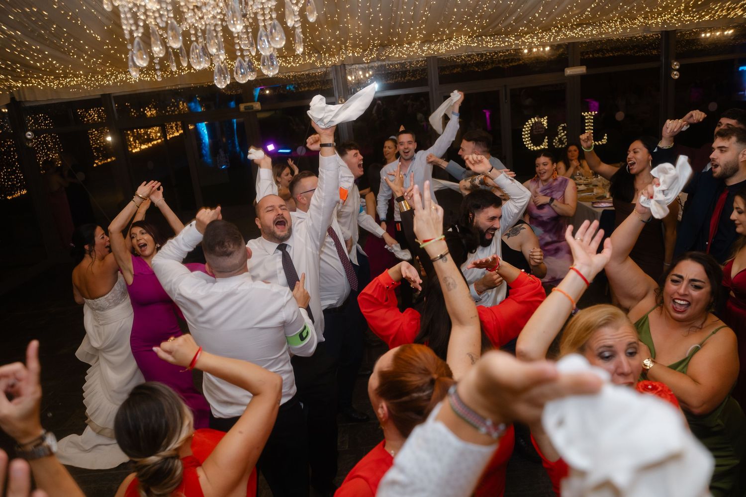 Large group of wedding guests dancing enthusiastically on a dimly lit reception dance floor.