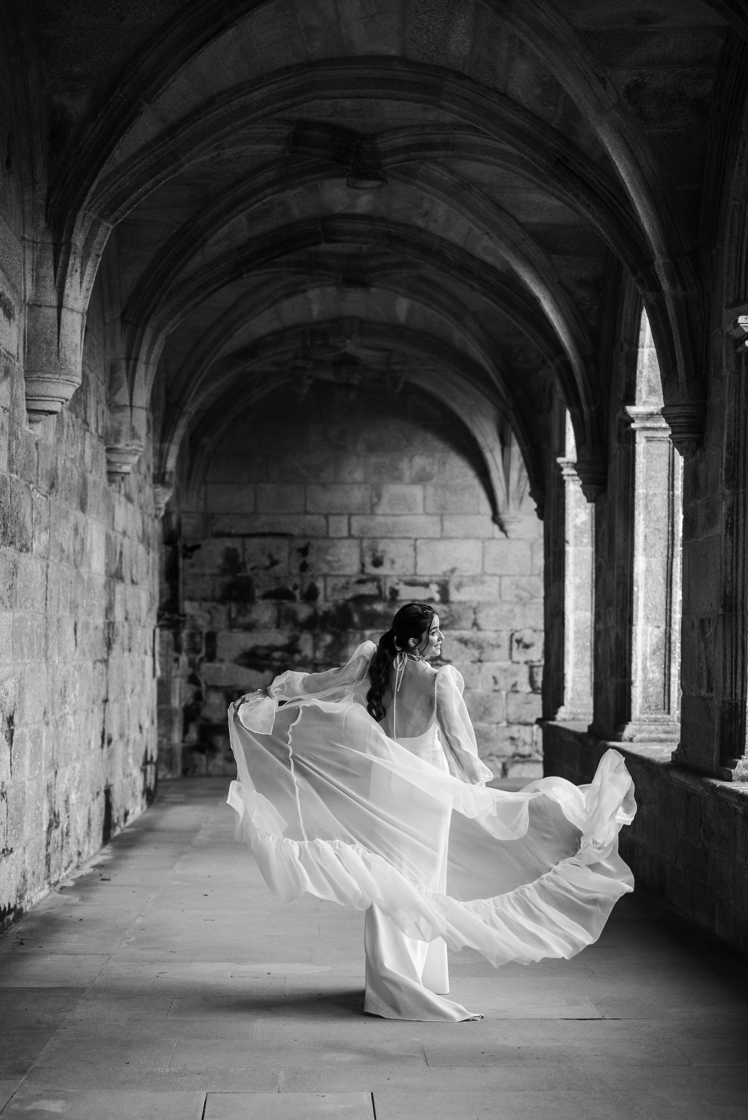 A flowing white fabric dances through a gothic stone archway corridor in a dramatic black and white photograph.