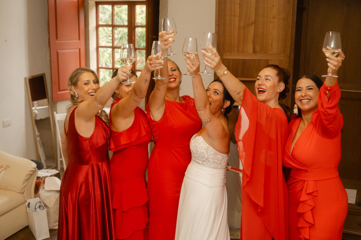 A bride in white and bridesmaids in bright coral dresses raise champagne glasses in celebration at a wedding.