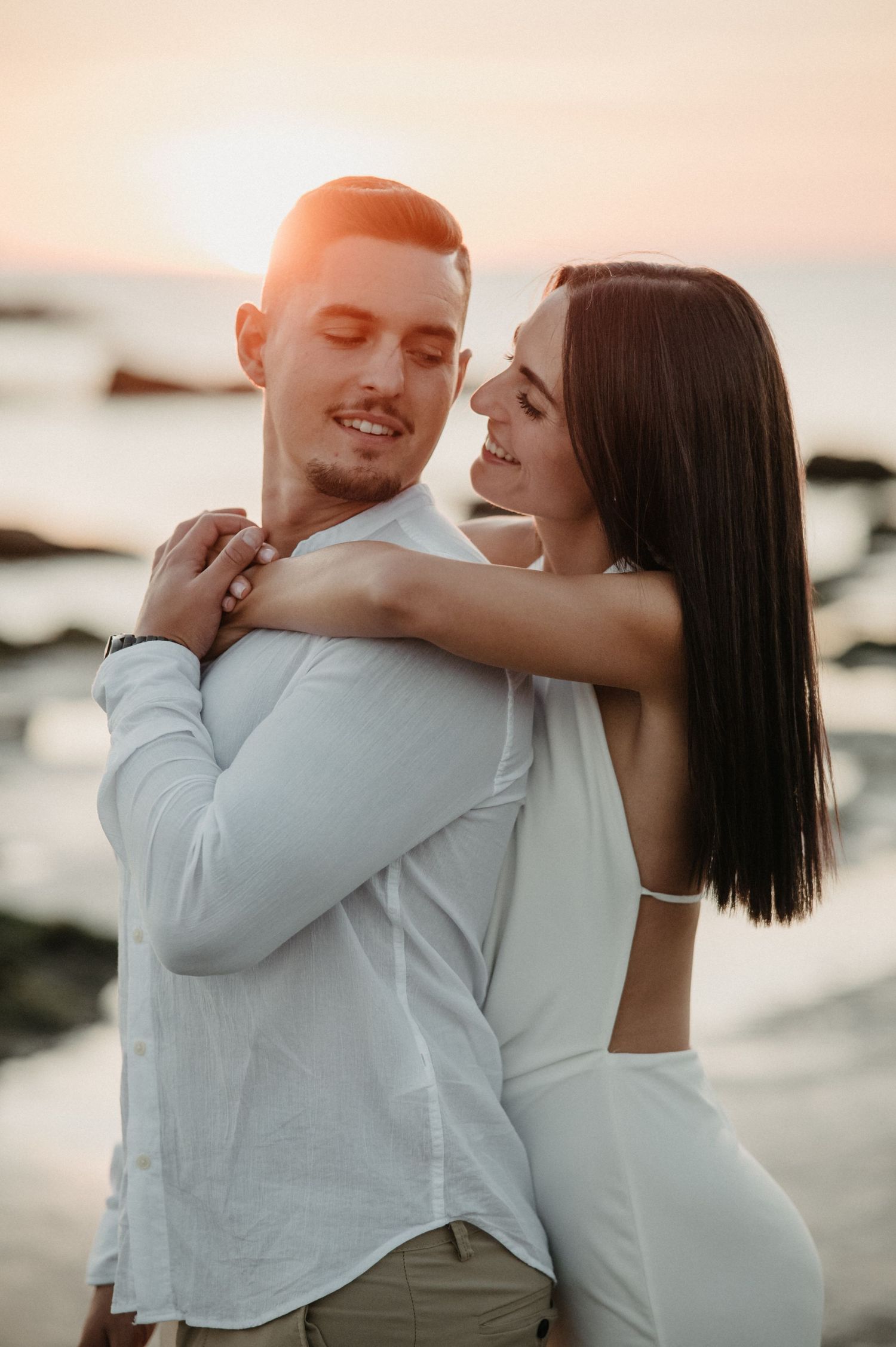 A couple embraces tenderly on a beach at sunset with soft pink lighting creating a romantic atmosphere.