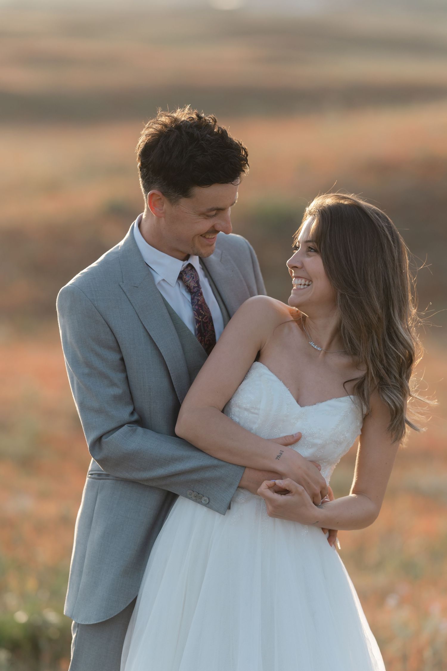 Happy bride and groom embracing and smiling at each other in a sun-kissed autumn field on wedding day.