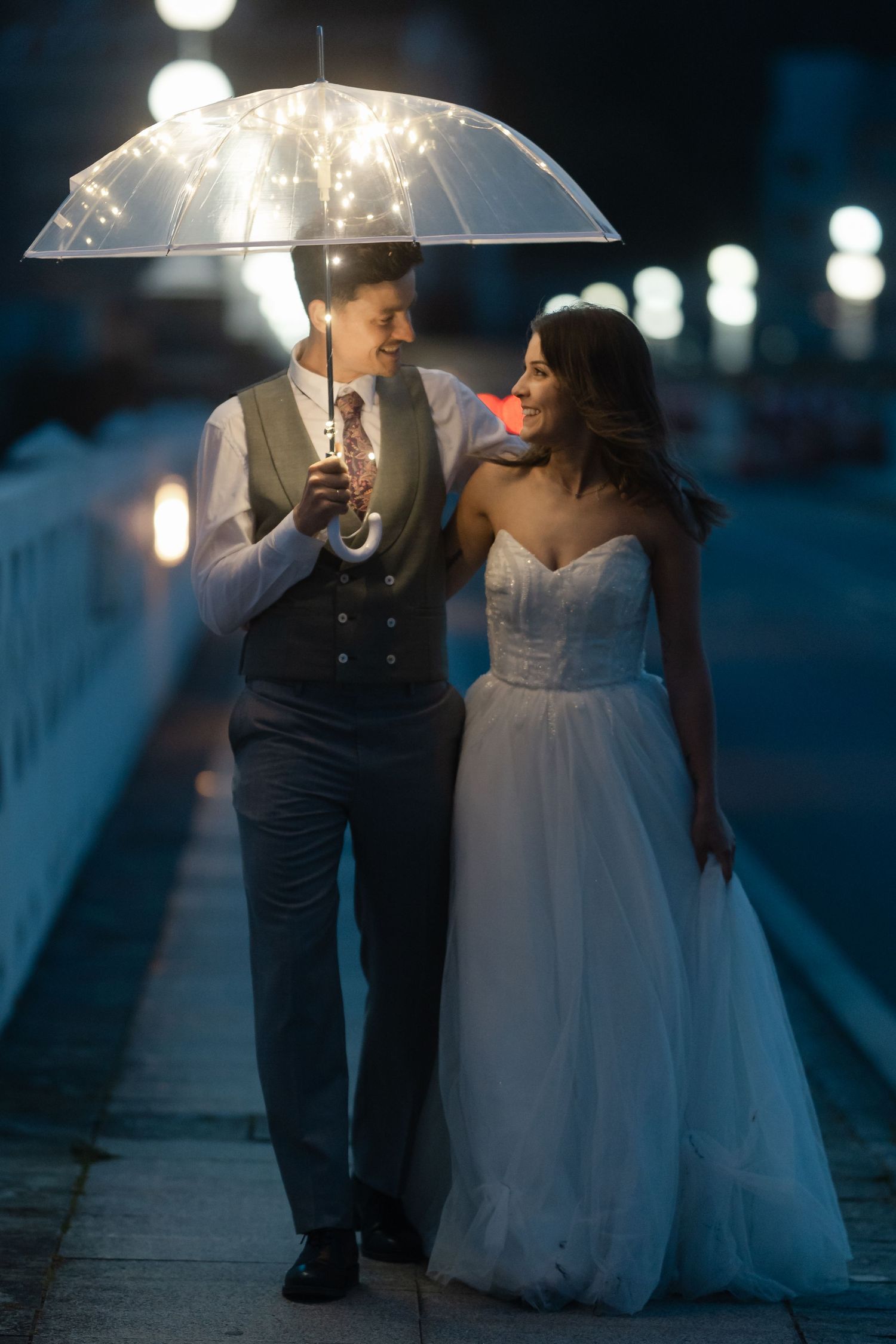 Bride and groom walking under a sparkling transparent umbrella at night, sharing a romantic glance.