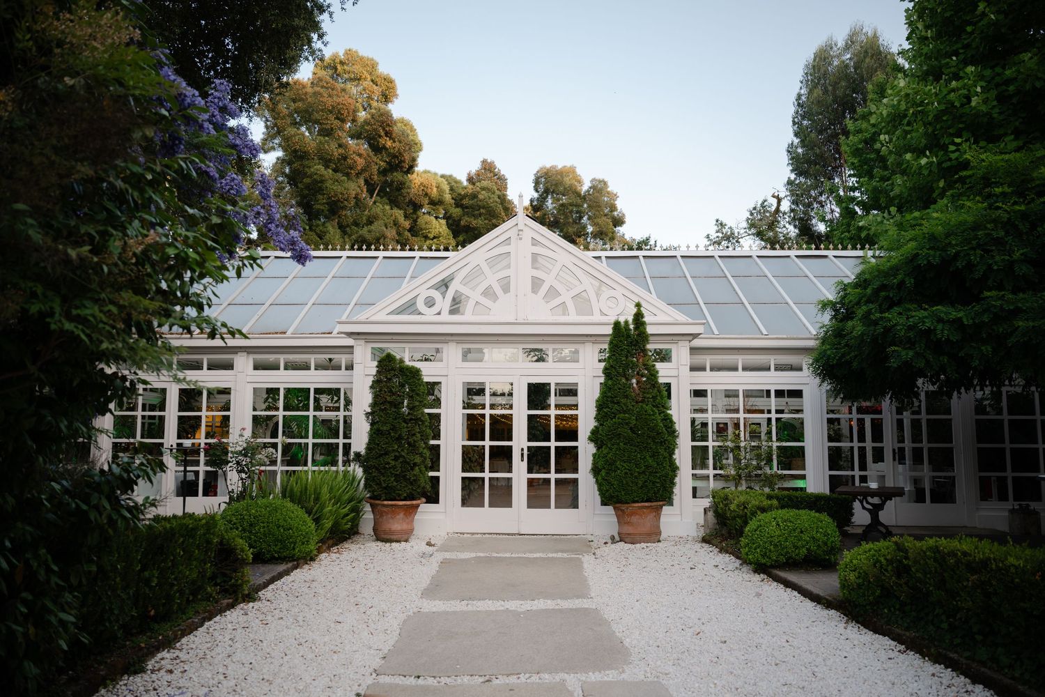 A series of connected white greenhouse conservatory buildings with glass panels and ornate architectural details in a garden setting.