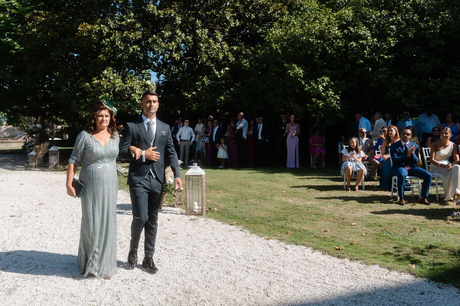 Wedding party walks along a white stone path surrounded by lush green trees and gardens.