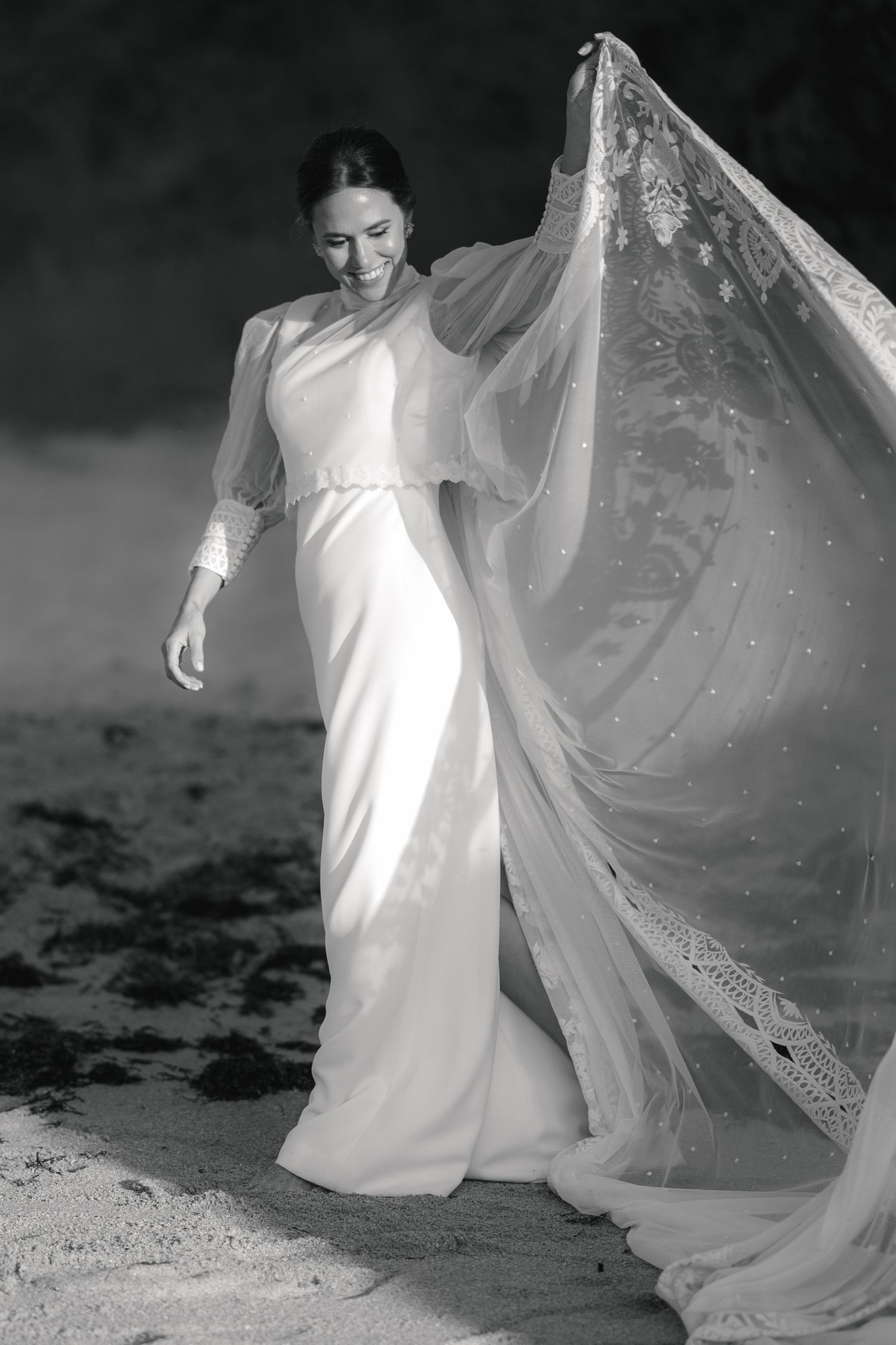 Bride in flowing white dress and veil twirls gracefully on a beach.