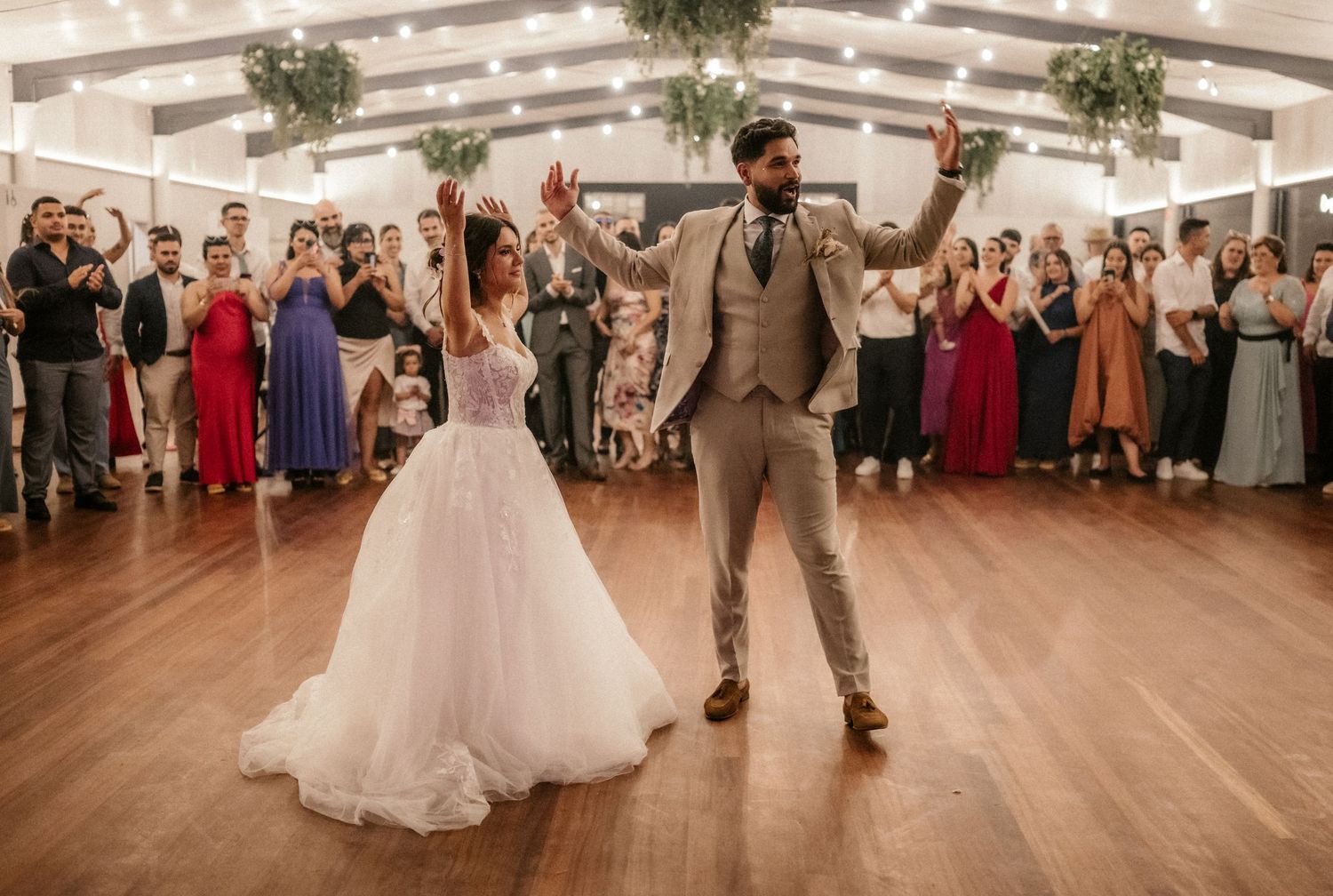 Wedding guests watch as dancers perform on a wooden dance floor under string lights and greenery.