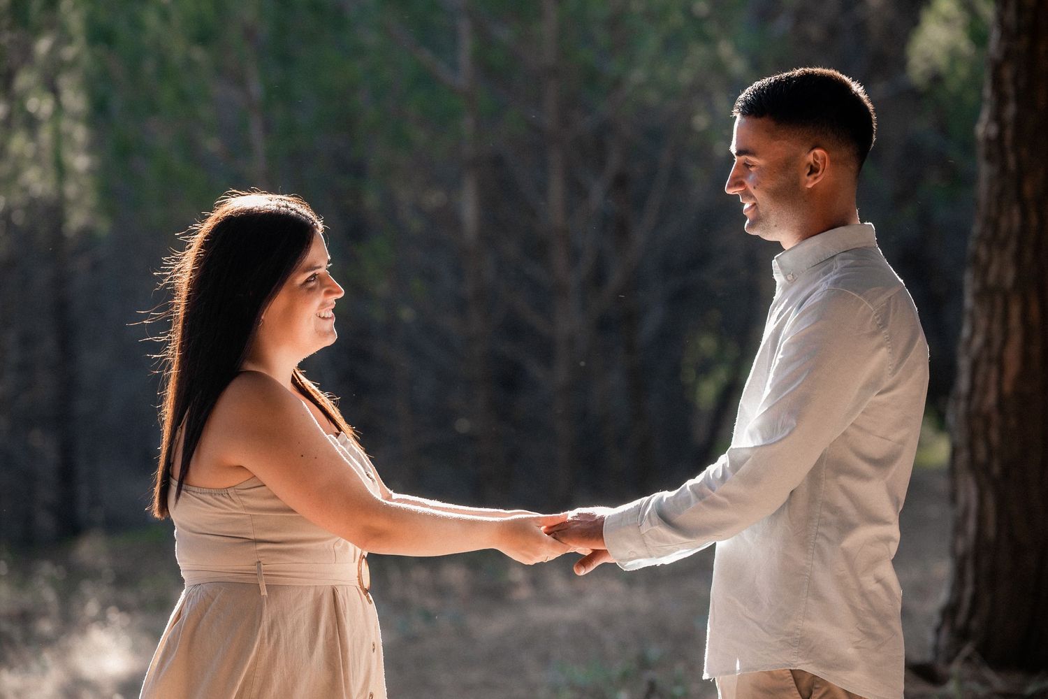 A couple in white outfits dance together in a sunlit forest setting during what appears to be an engagement photoshoot.