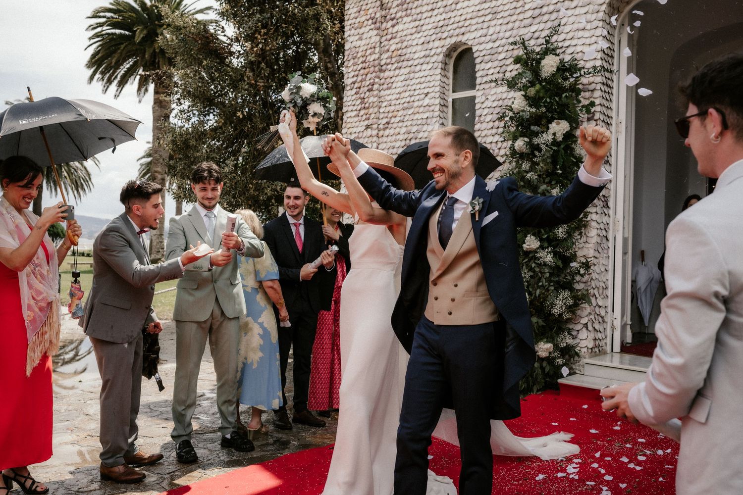 Joyful wedding celebration with guests tossing rose petals on a red carpet outside a stone building.