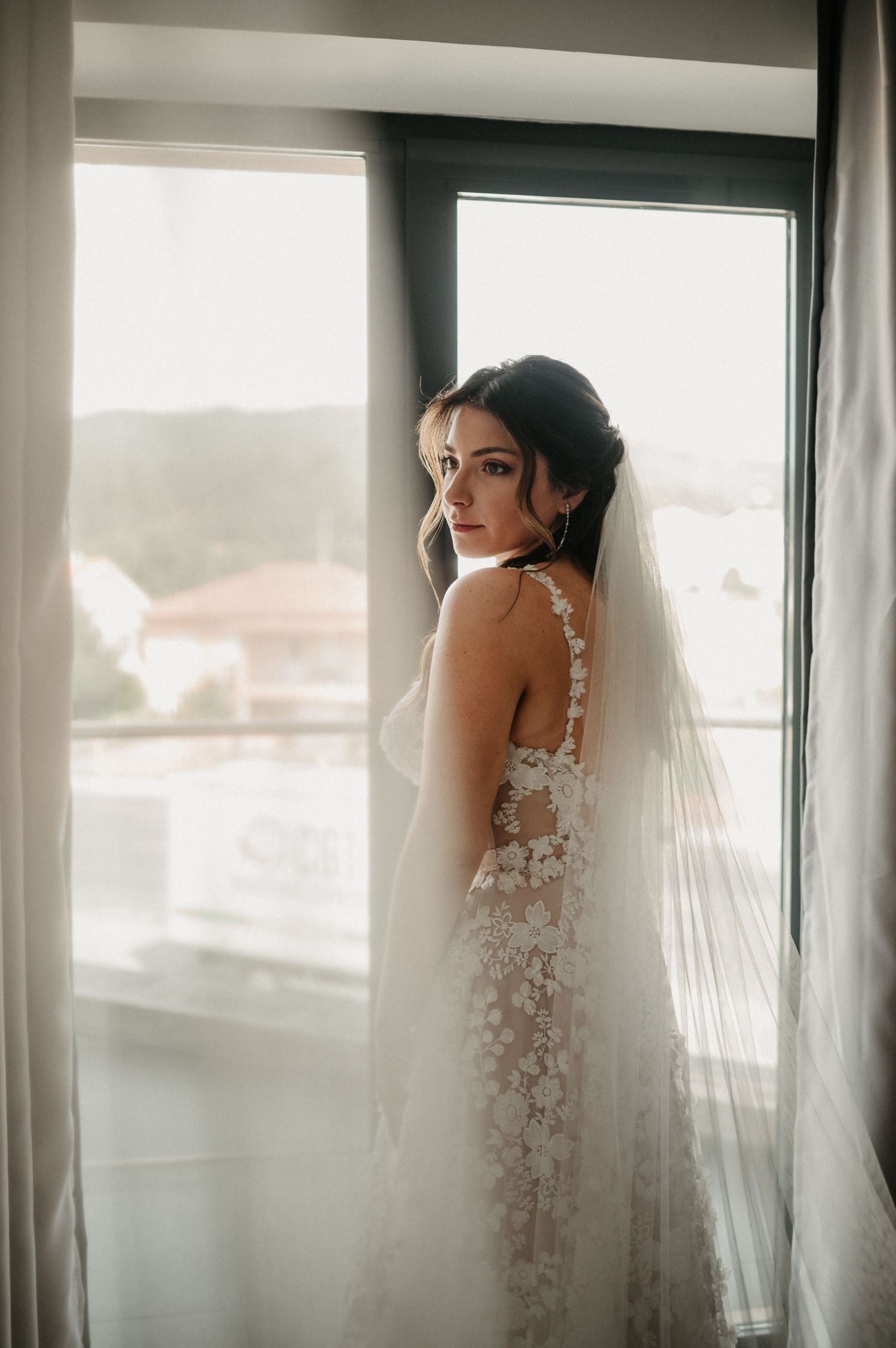 Bride in delicate lace wedding dress stands near sunlit windows in artistic bridal portrait series.